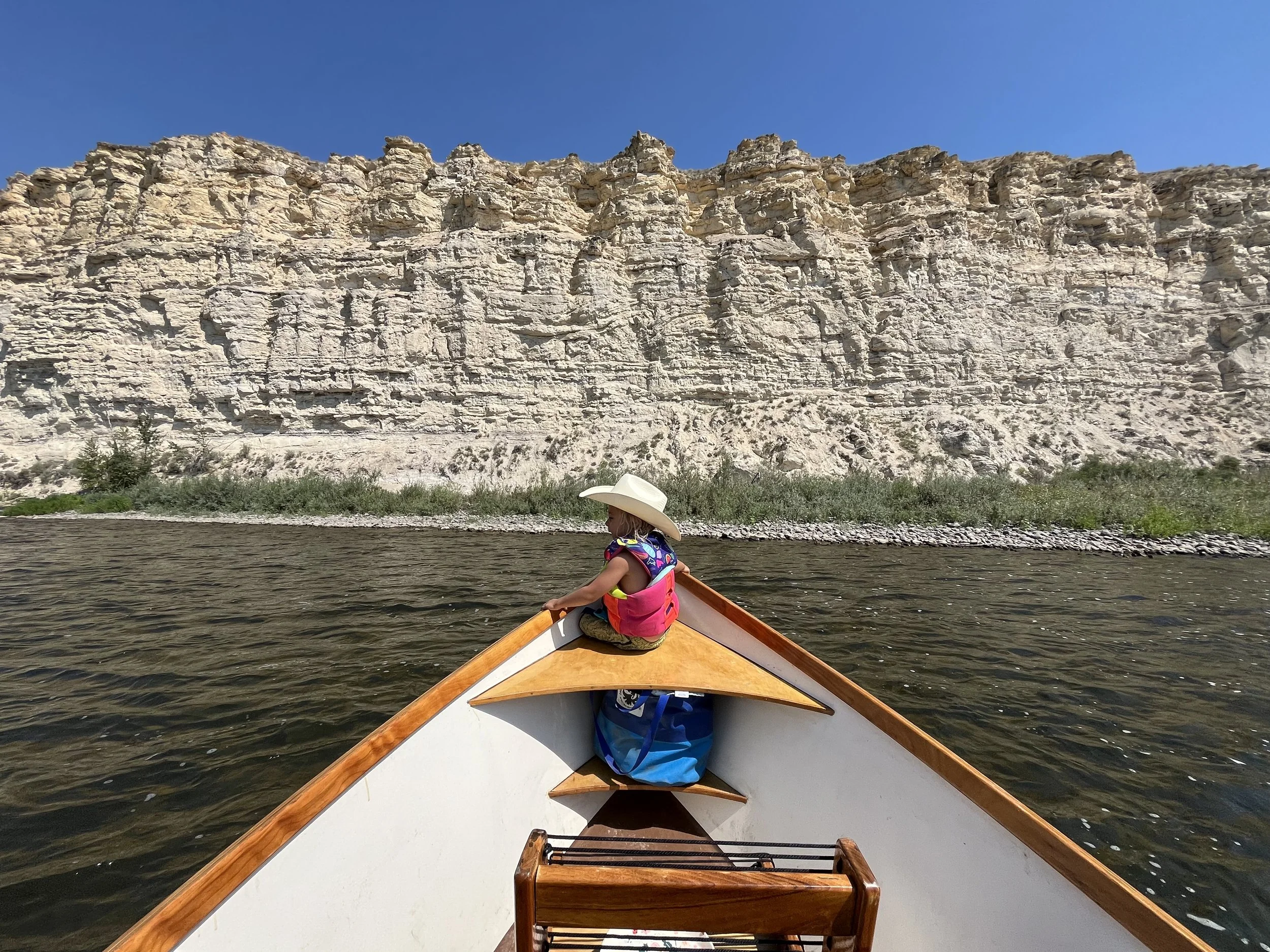 A young girl wearing a sun hat and life vest paddling a canoe on a river, with a large rocky cliff in the background under a clear blue sky.