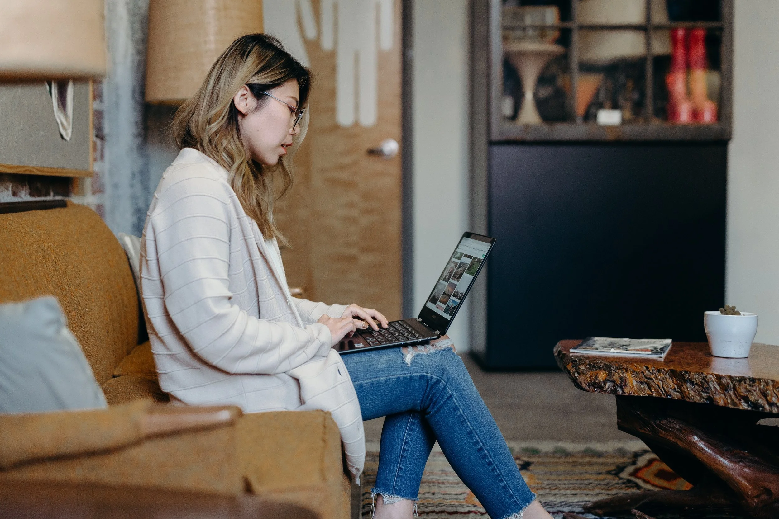 A young woman with glasses and shoulder-length hair is sitting on a brown sofa, working on a laptop in a cozy living room with wooden and brick decor.