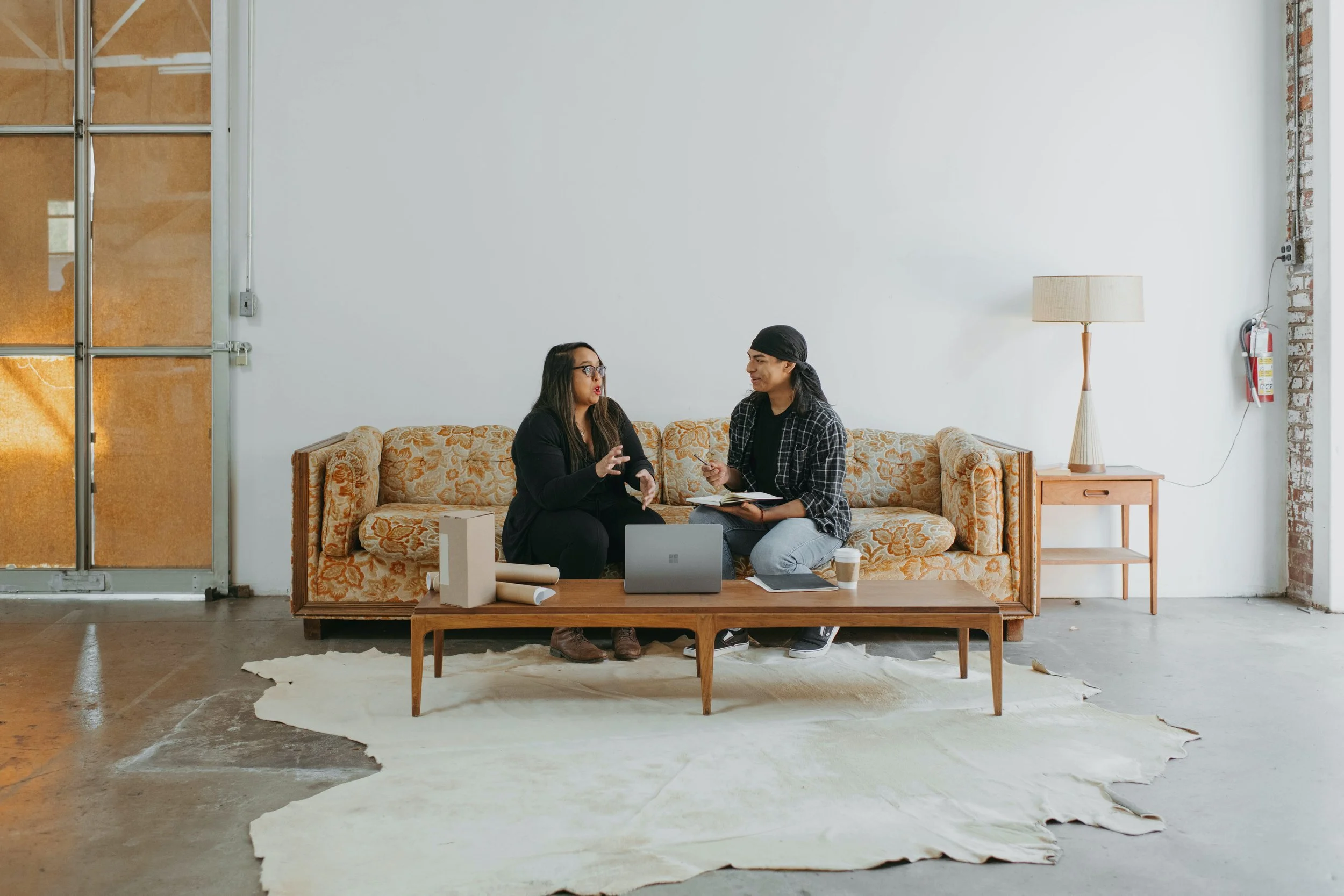 Two women sitting on a patterned beige sofa in a casual setting, engaged in conversation. One woman with glasses and long dark hair gesturing, the other with long dark hair and a black headscarf, holding a notebook and pen. A laptop, coffee cup, and cardboard tubes are on the wooden coffee table in front of them, with a white rug on a concrete floor. The background includes a plain white wall, a wooden side table with a lamp, and an outlet with a fire extinguisher nearby.