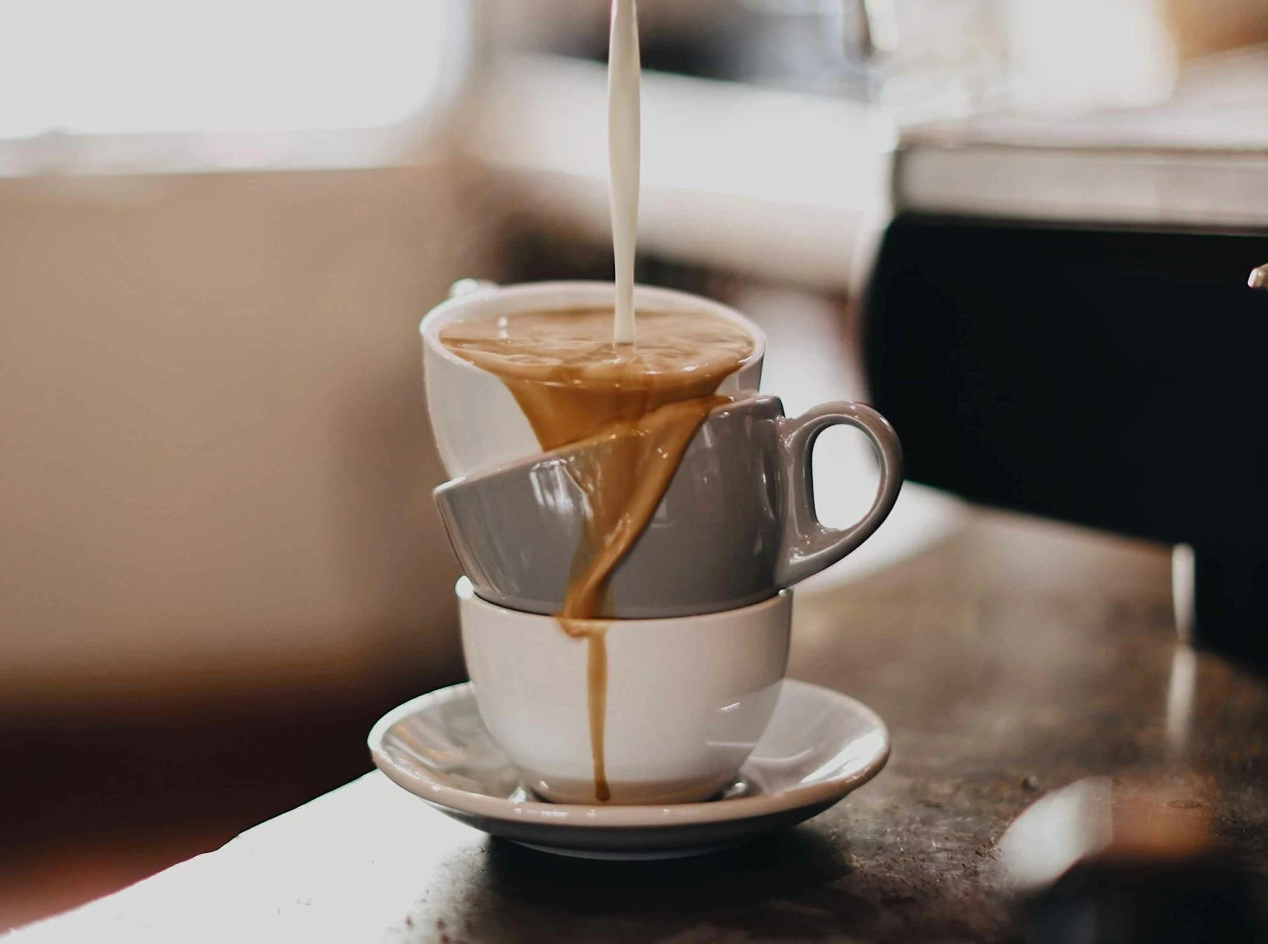 Milk being poured into a stack of three coffee cups on a wooden table.