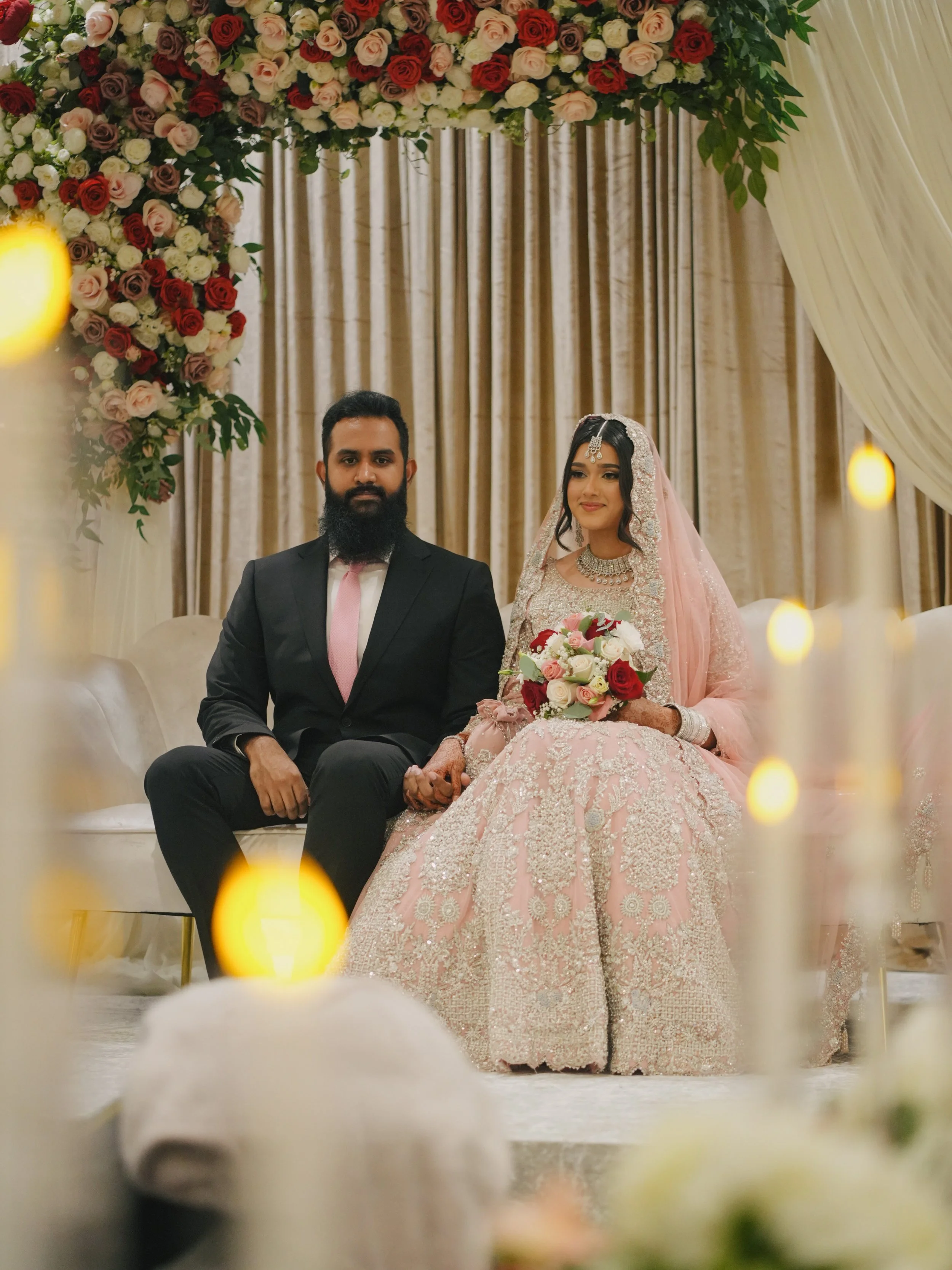 A couple dressed in wedding attire sitting on a sofa, holding hands, in front of a backdrop adorned with a floral arrangement of red, pink, and white roses, with beige curtains.