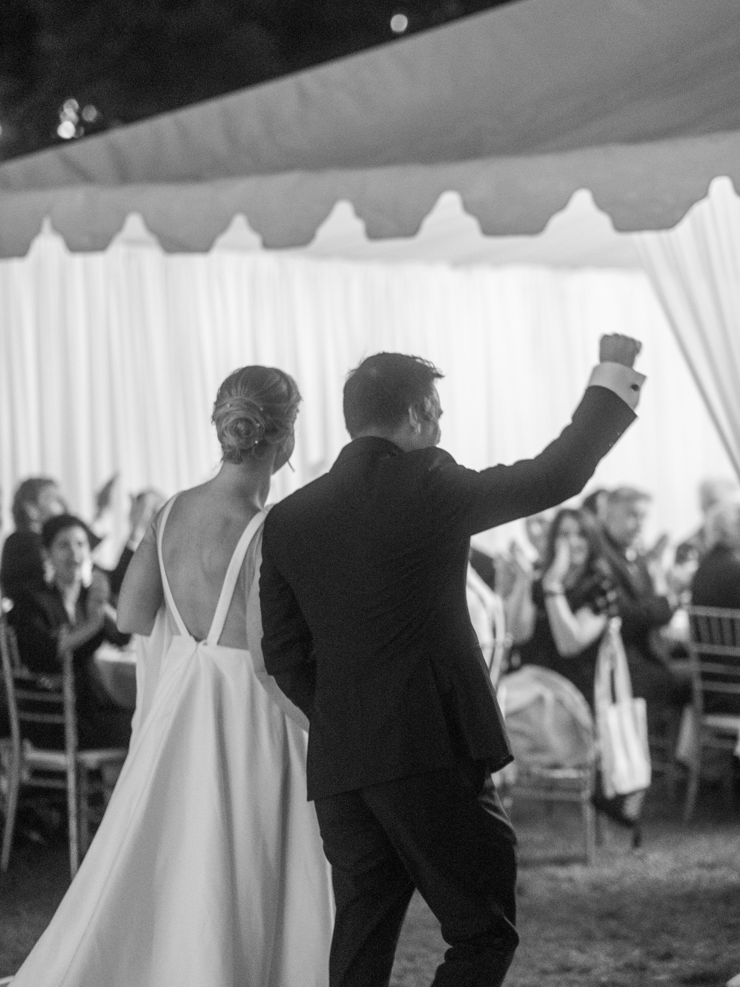 A black and white photo of a wedding reception. A bride and groom are dancing; the groom has his fist raised in celebration, while the bride looks on. Guests sit at tables in the background, watching the couple.