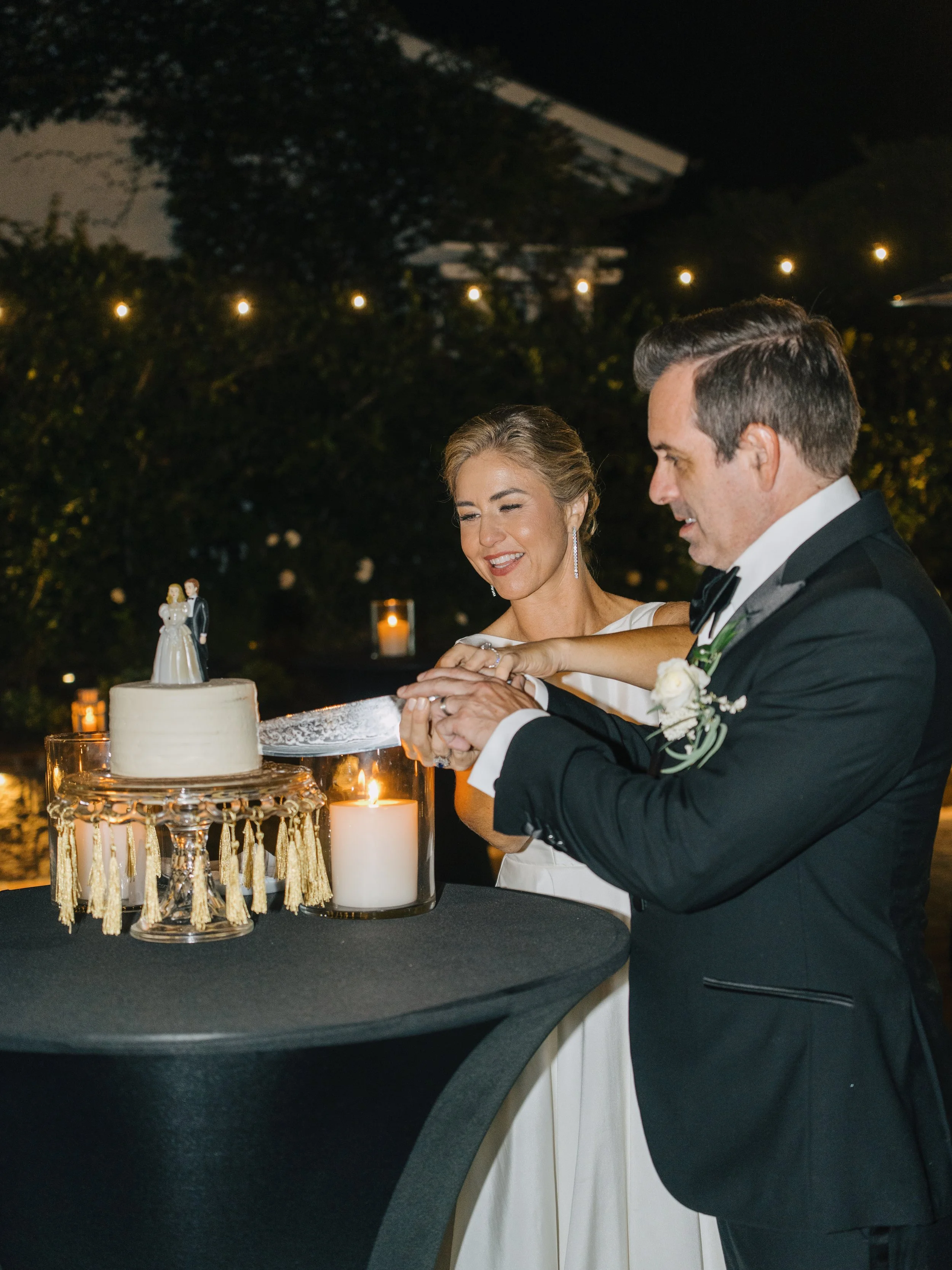 A bride and groom at their wedding reception cutting a wedding cake together, outdoors at night with candles and string lights.