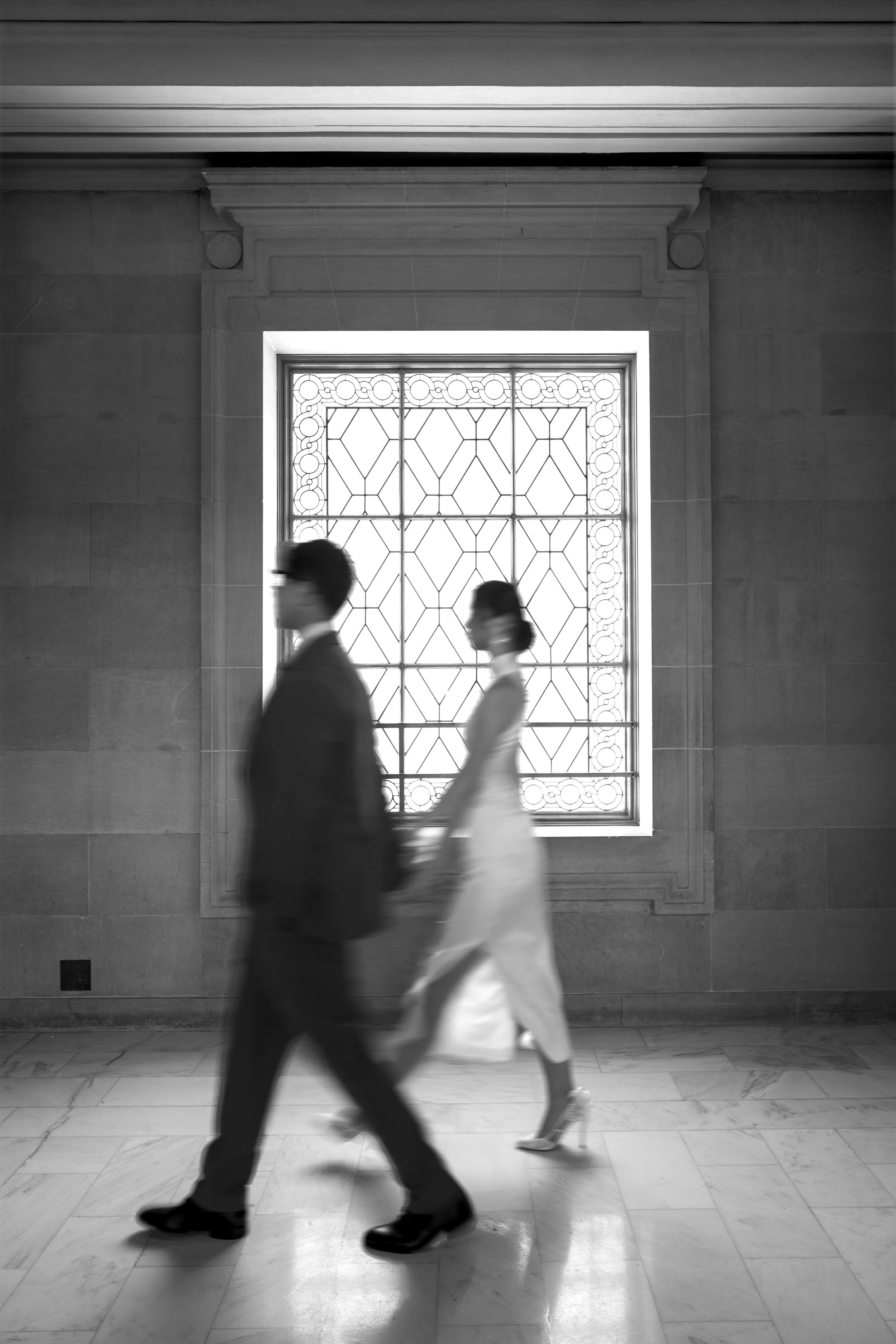 Black and white photo of a man and woman walking past a large ornate window. The man is dressed in a suit and wearing glasses; the woman is in a dress and high heels. The window has decorative metalwork and allows light to come in, illuminating the marble floor.