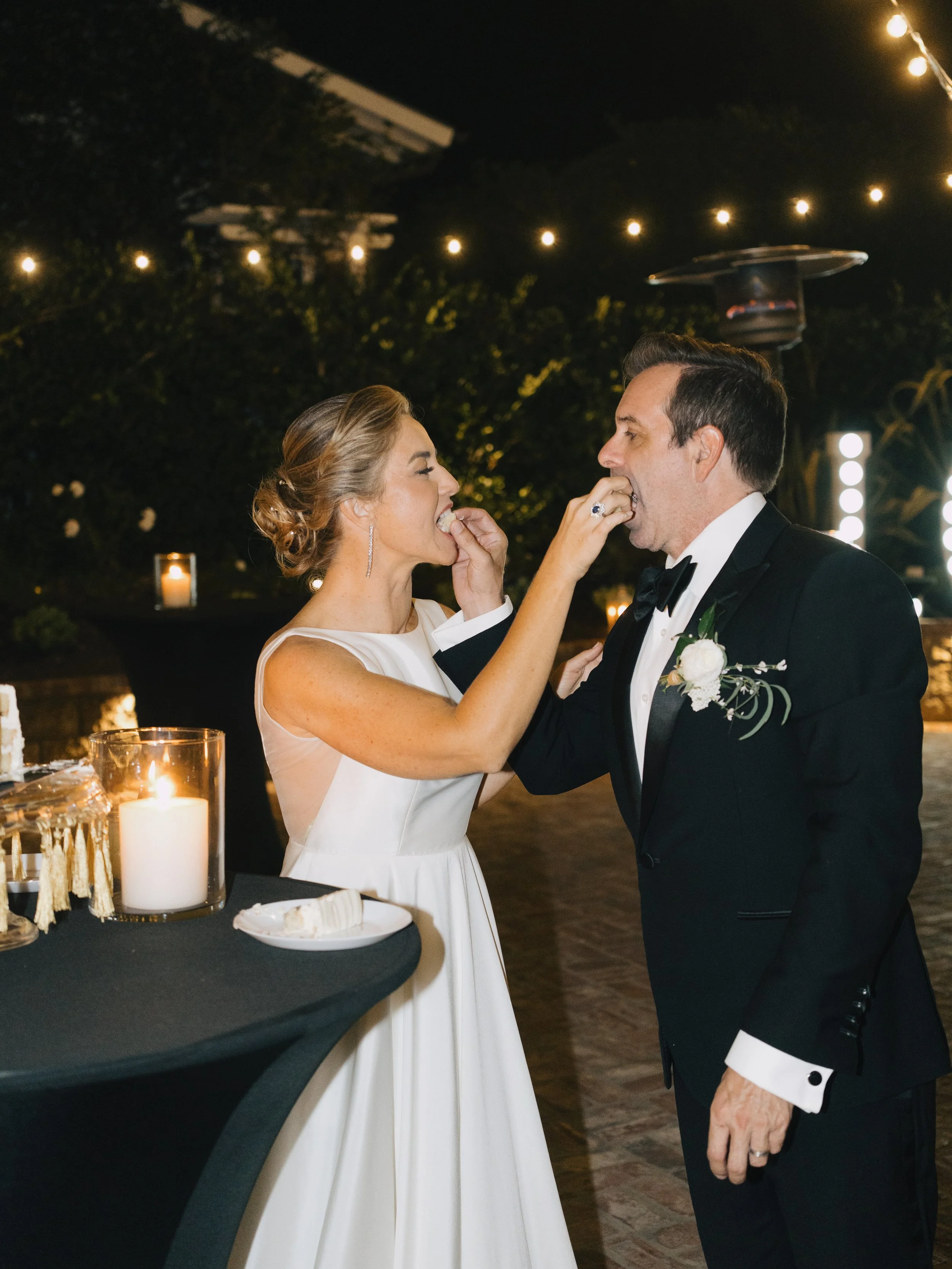 A bride and groom feed each other cake at their wedding reception outdoors at night, decorated with string lights and candles.