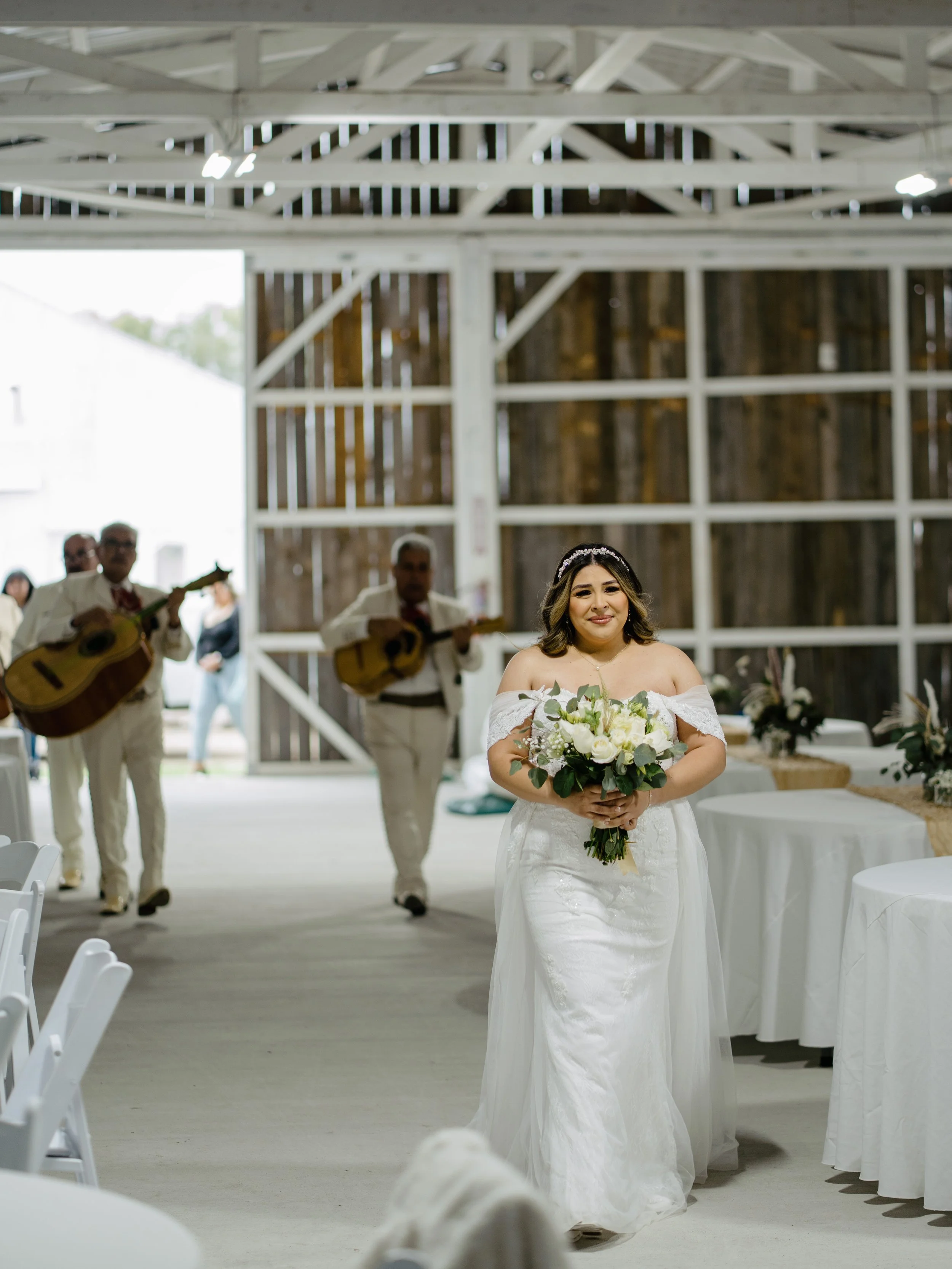 A bride in a white off-the-shoulder wedding dress holding a bouquet of white and green flowers, walking inside a rustic barn with guitar-playing musicians in the background.