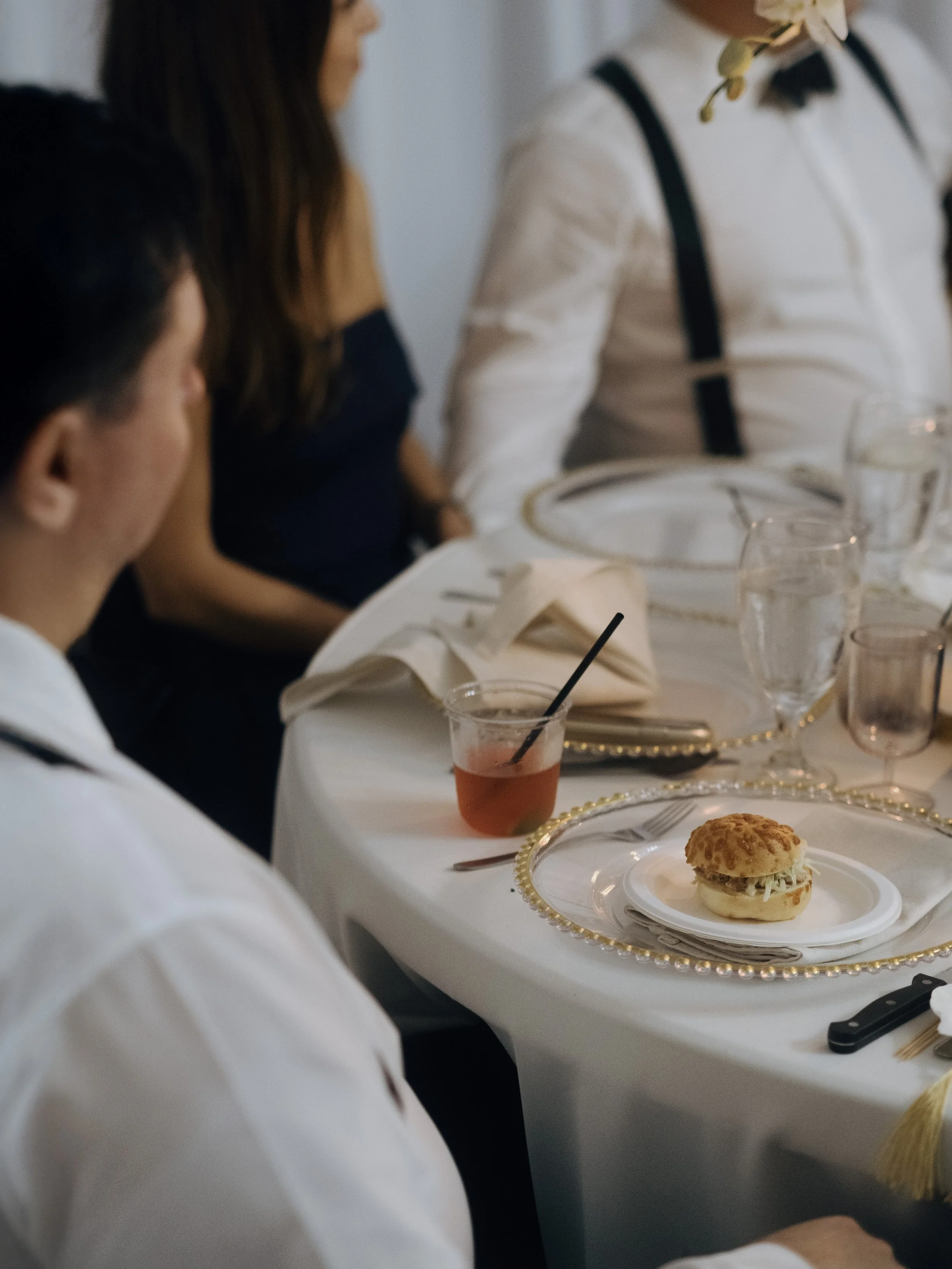 A dining table set for a formal event with plates, glasses, and napkins. There is a small sandwich on a decorative plate, a drink with a straw, and other glassware on the table. People are seated around the table.