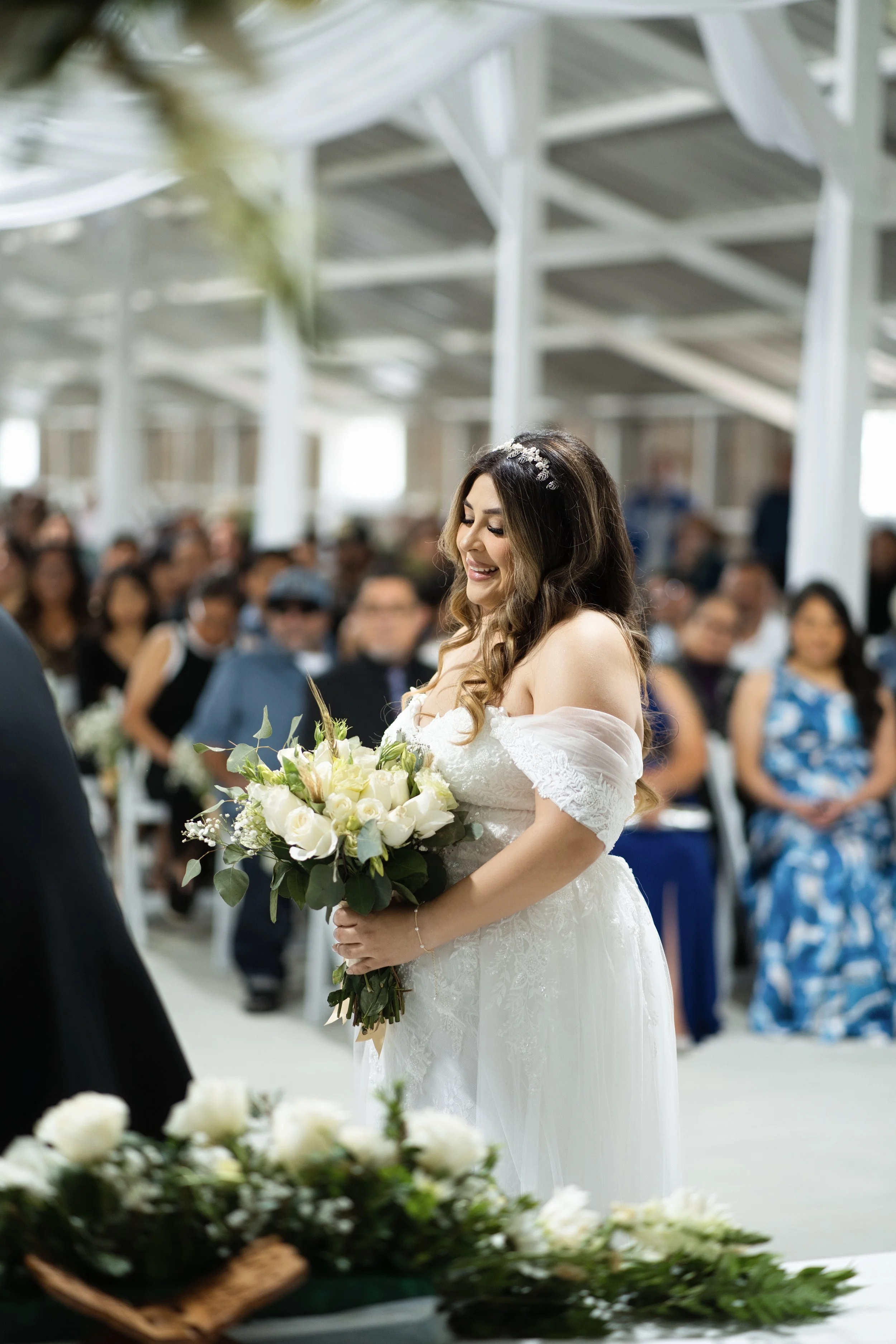 A bride with long, wavy brown hair wearing an off-shoulder white wedding dress holding a bouquet of white roses, standing inside a bright, indoor wedding venue with guests in the background.