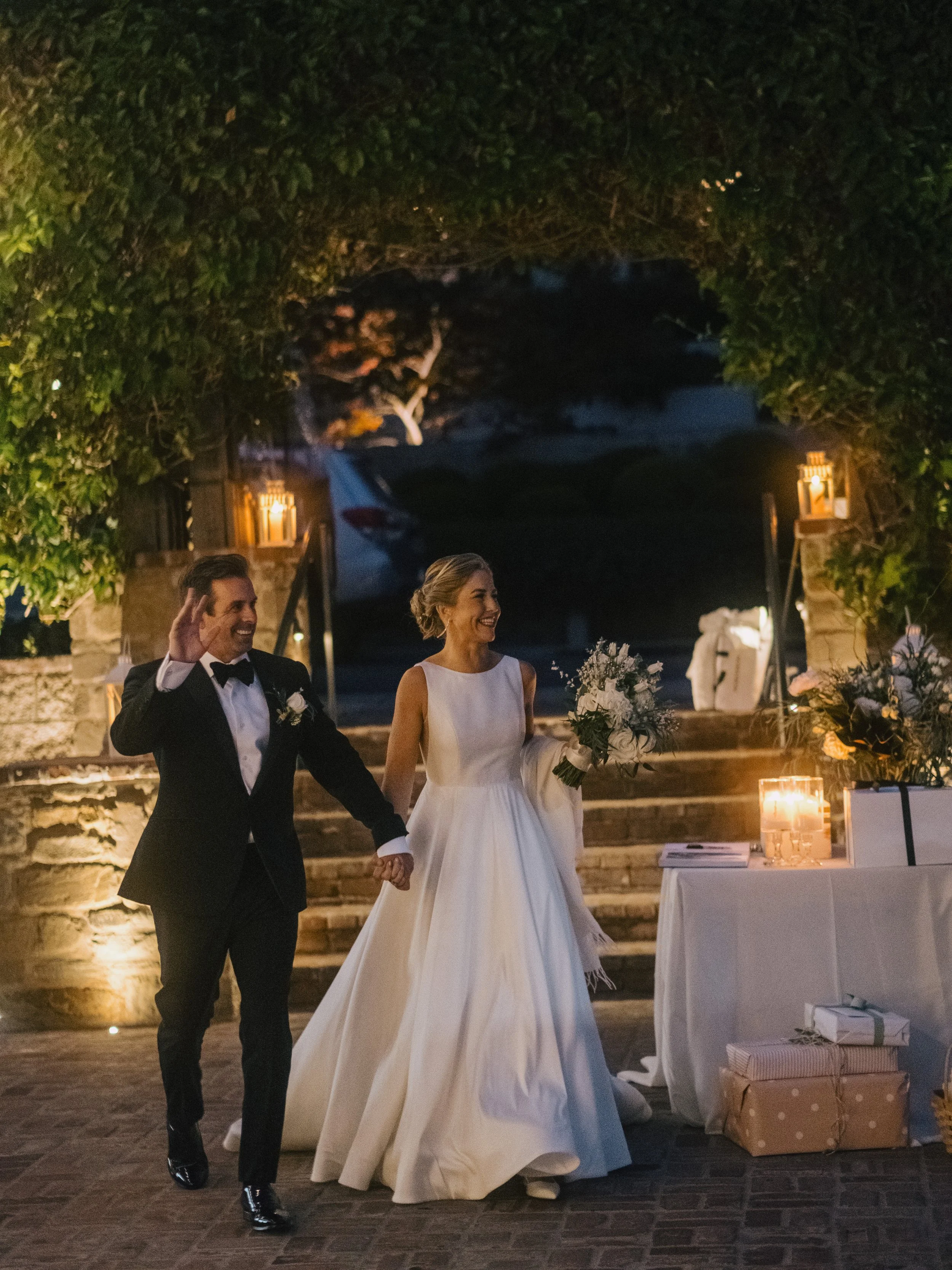 A bride and groom walking hand in hand at their wedding reception outdoors at night. The groom is wearing a black tuxedo and the bride is in a long white wedding gown holding a bouquet of white flowers. They are smiling and appear happy.