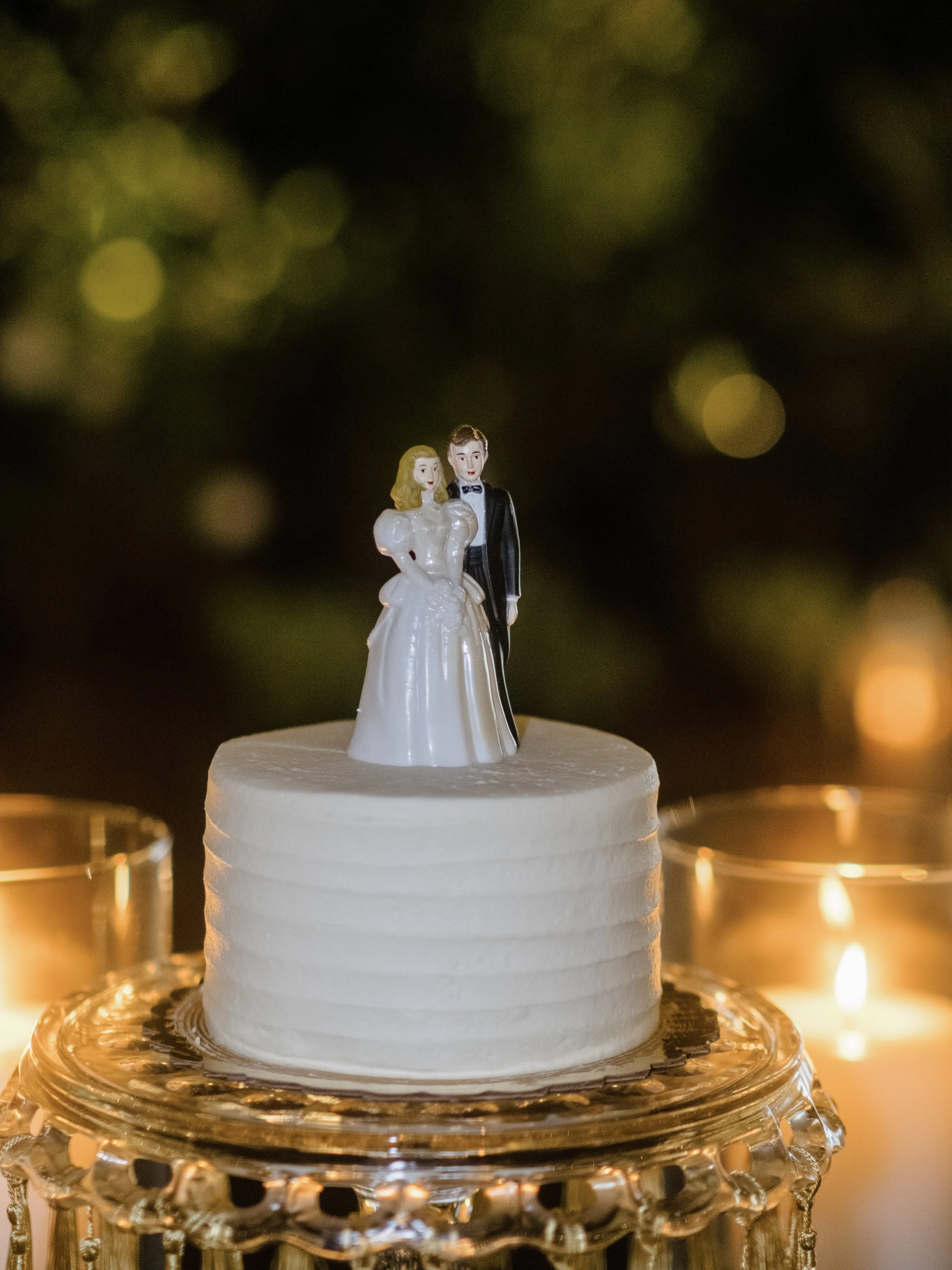 Small wedding cake with a bride and groom figurine on top, placed on a decorative stand surrounded by lit candles.