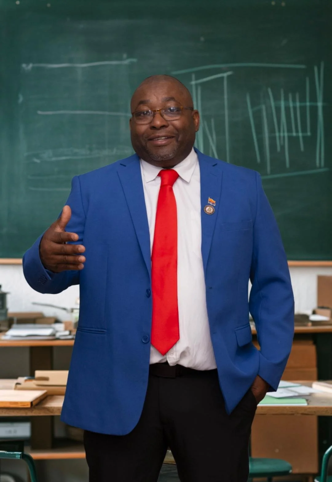 A man in a blue suit, red tie, and white shirt standing in front of a chalkboard in a classroom, gesturing with his right hand.