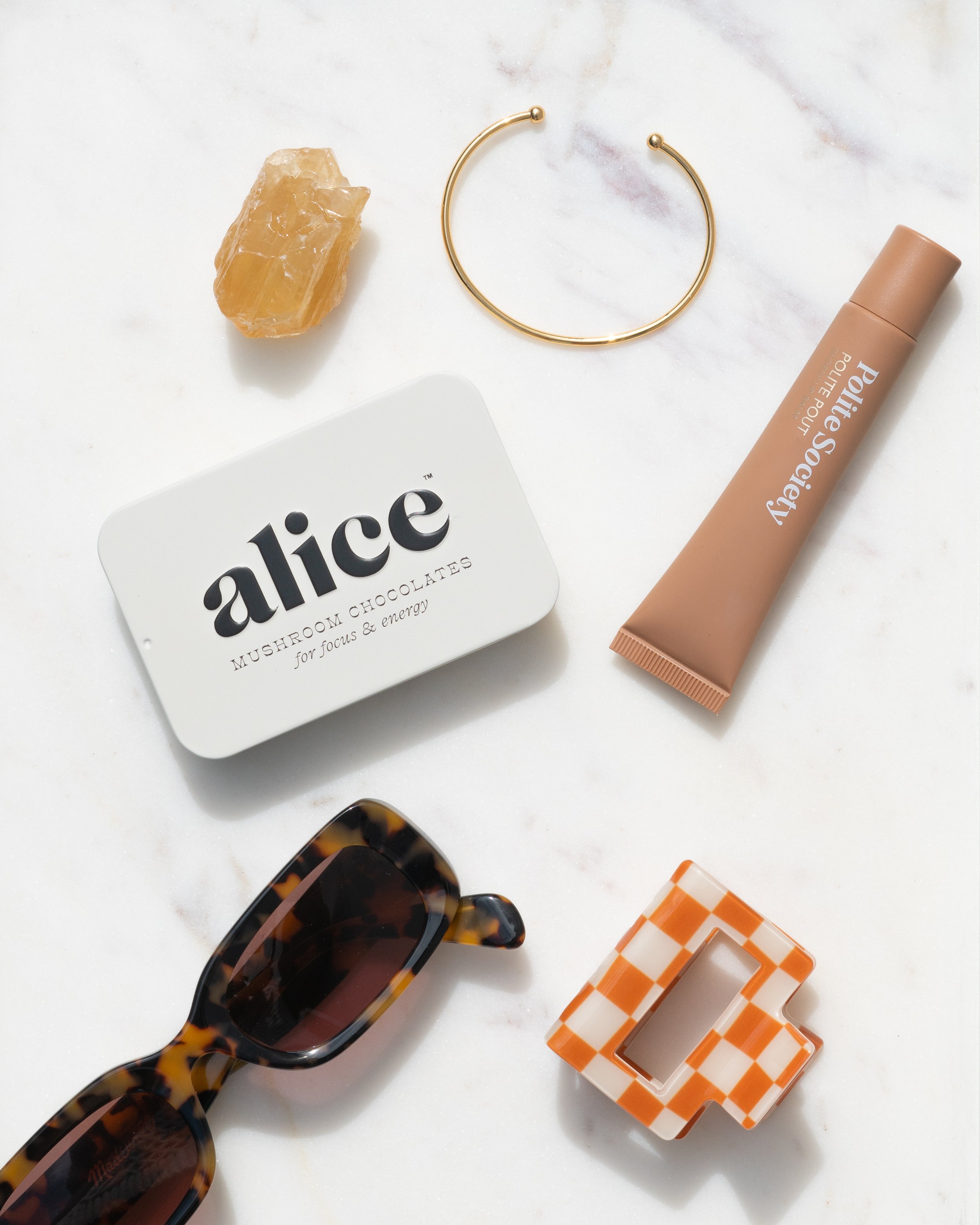 Flat lay photography of a gold cuff bracelet, amber crystal, nude lipstick, mushroom chocolate tin, tortoiseshell sunglasses, and orange-and-white checkered hair clip on white marble.