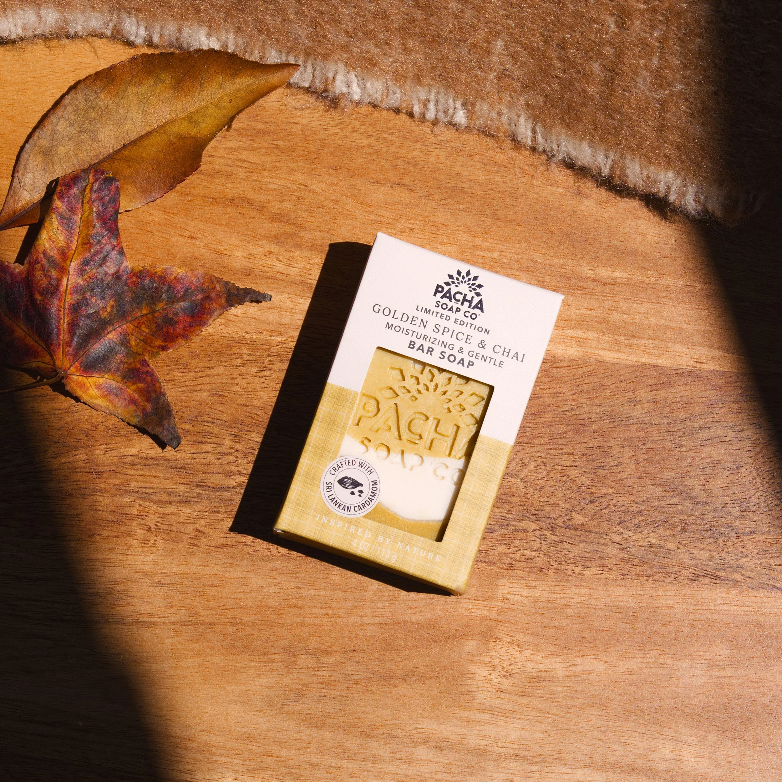 Elevated flat lay product shot of a Golden Spice & Chai moisturizing bar soap in packaging on a wooden table with autumn leaves and a cozy knit blanket.