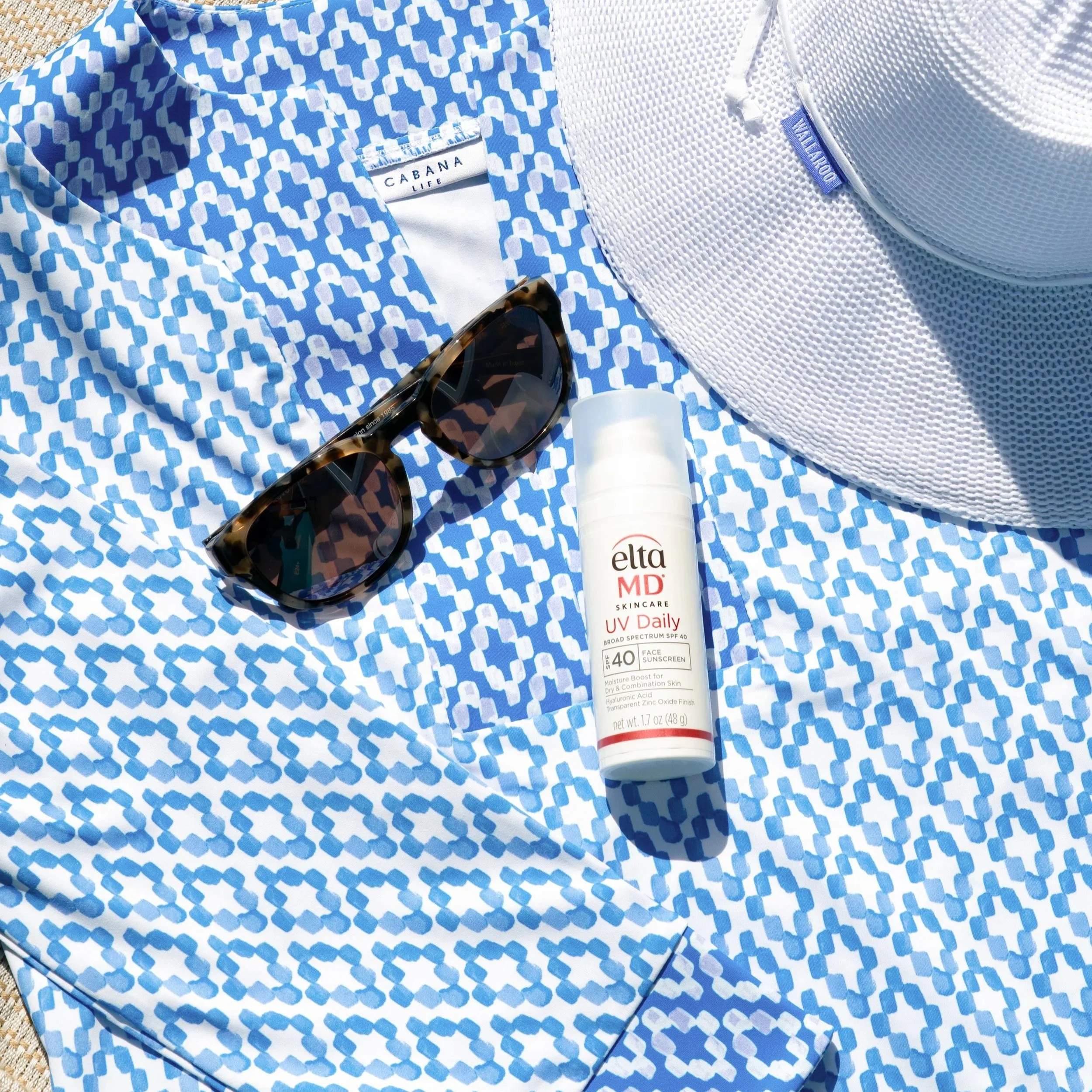 Beachwear clothing photography, sunglasses, a white sun hat, and a bottle of EltaMD UV daily sunscreen on a towel, flatlay product photographer Los Angeles