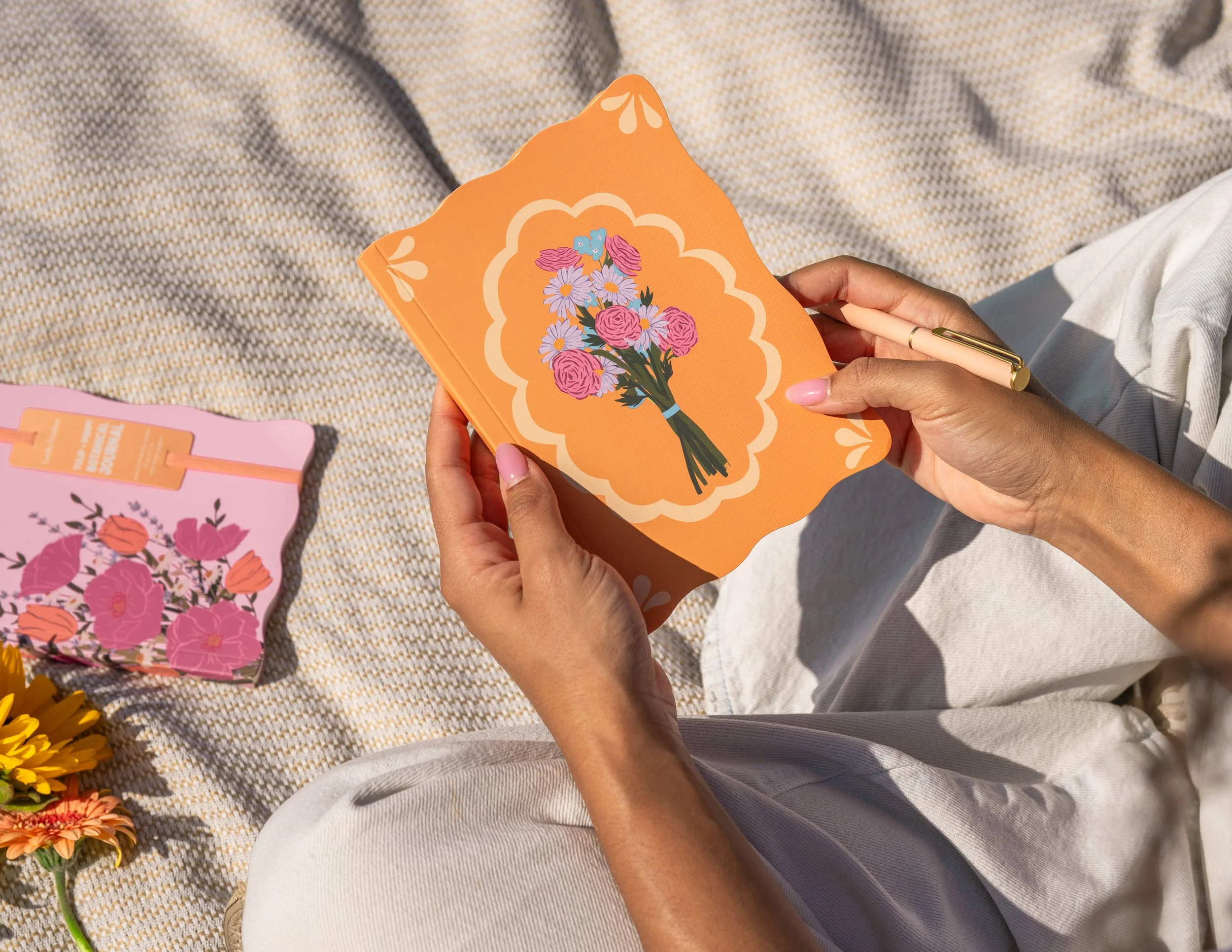 Elevated outdoor lifestyle photograph of a hand model holding an orange floral notebook and pen while sitting on a textured blanket surrounded by flowers.
