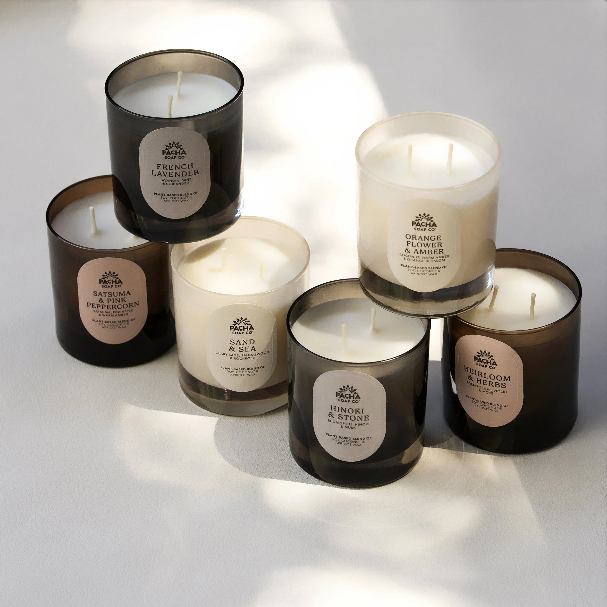 Clean studio product photography of a collection of Pacha Soap Co. scented candles in black, white, and amber glass jars, including French Lavender, Orange Flower & Amber, and Hinoki & Stone.