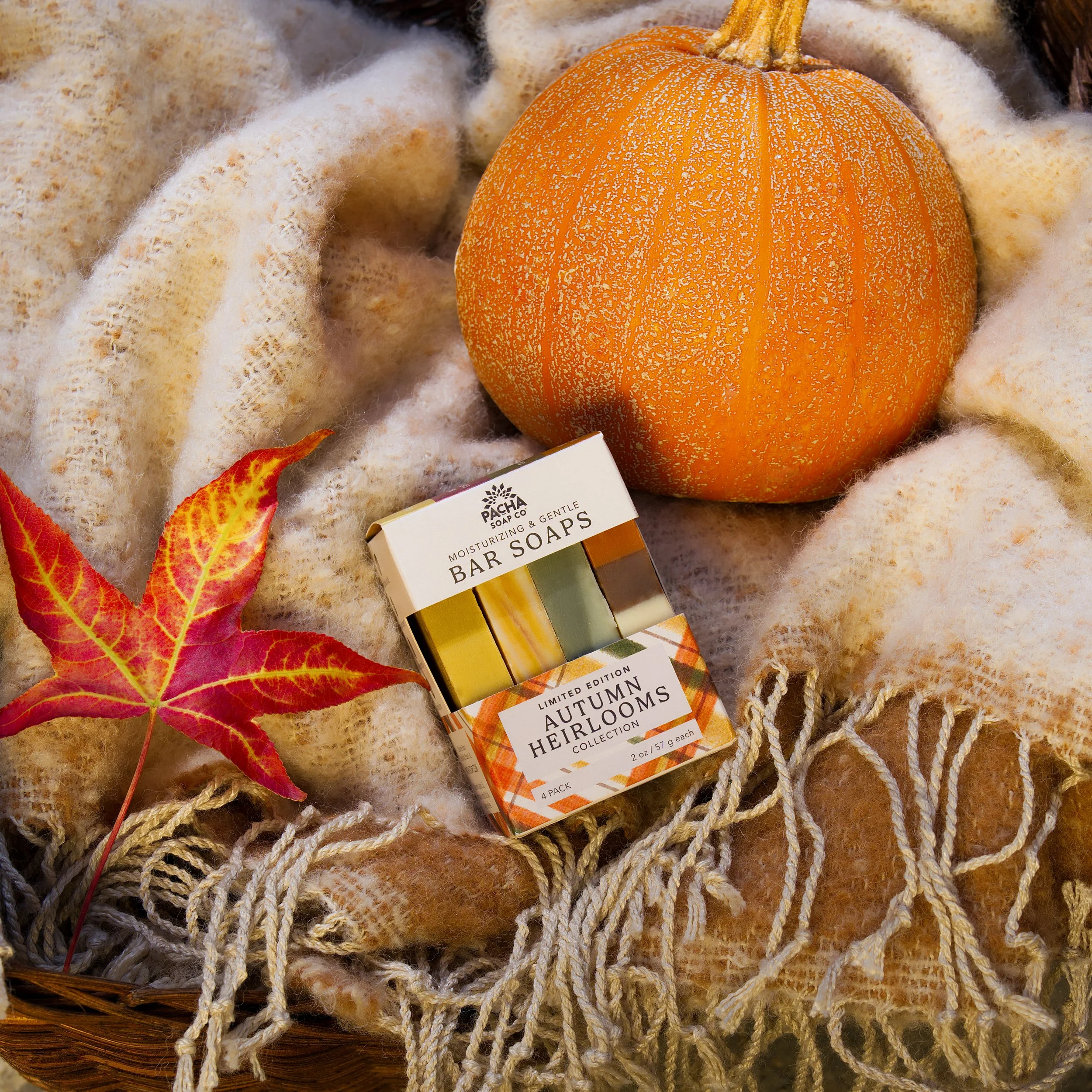 Handmade soap product photography of an autumn bar soap gift set beside an orange pumpkin, red autumn leaves, and rustic woven knit blanket.