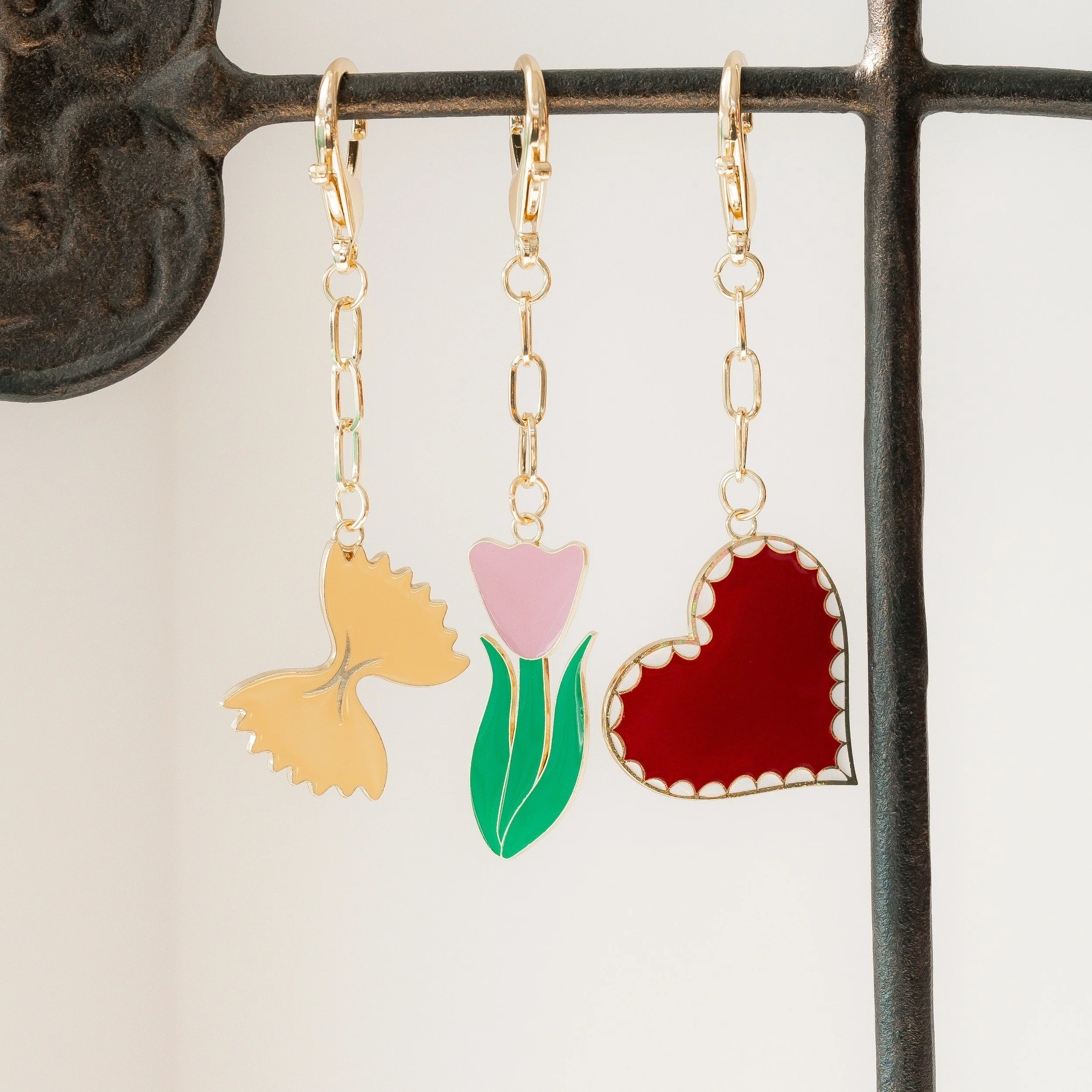 Catalog product photography of gold bag charms of a ravioli, tulip, and red heart hanging from a brass jewelry stand.
