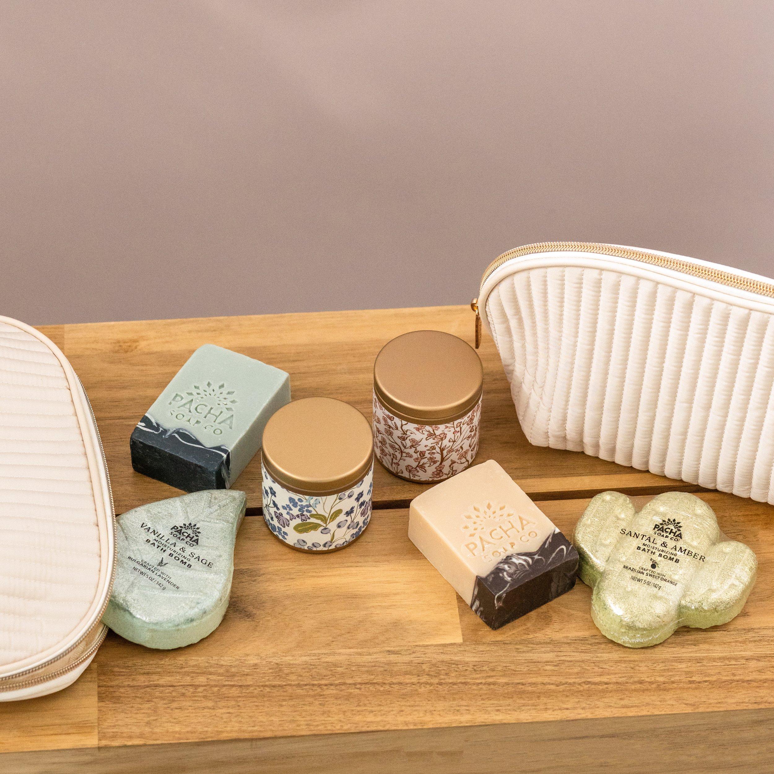 Elevated group product photography in a clean studio setting of bath soaps, candles, and bath bombs in cream travel bags on a wooden table.
