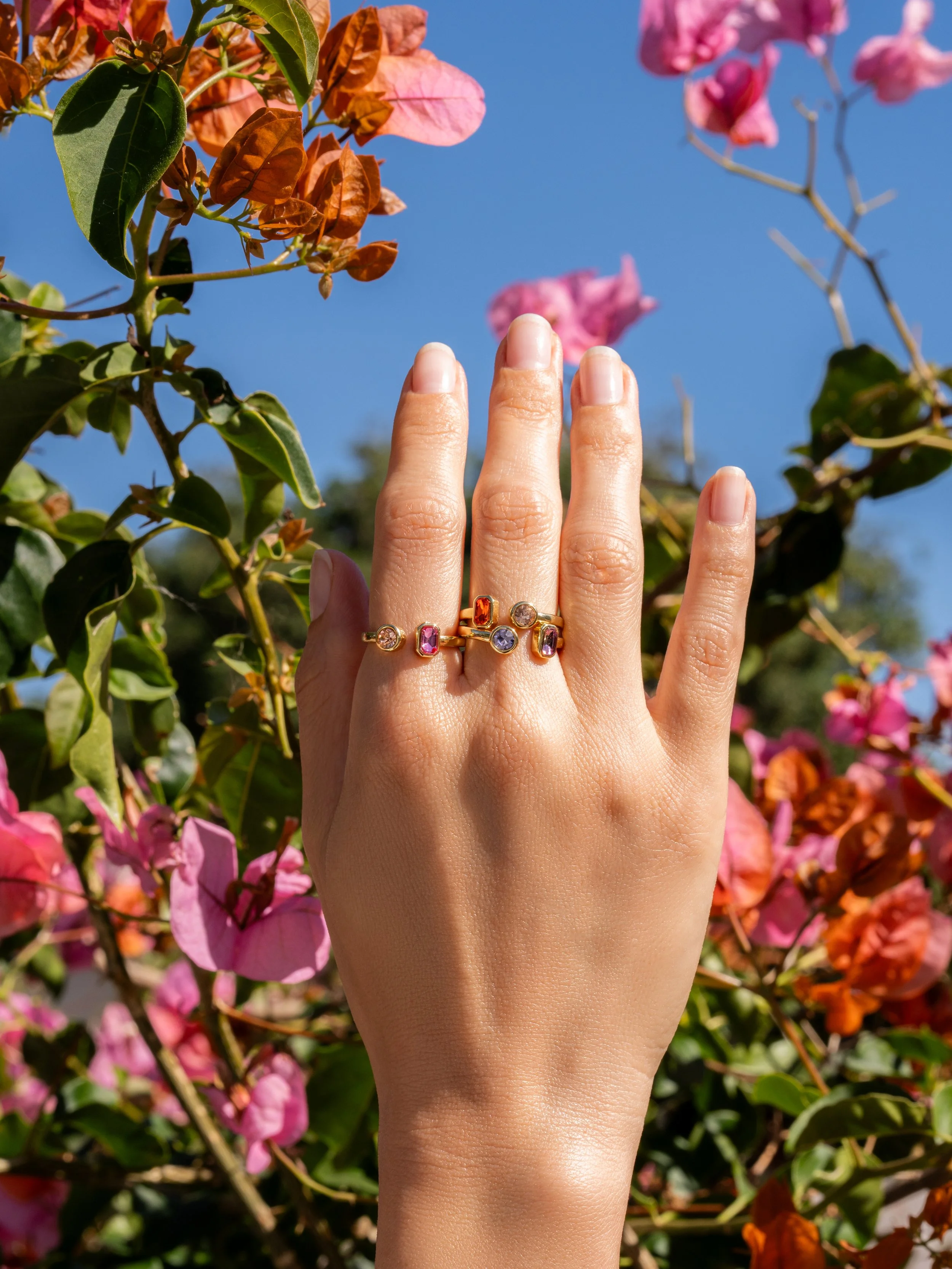 Elevated outdoor lifestyle photography of a hand model wearing colorful gemstone rings in front of pink and orange bougainvillea flowers.