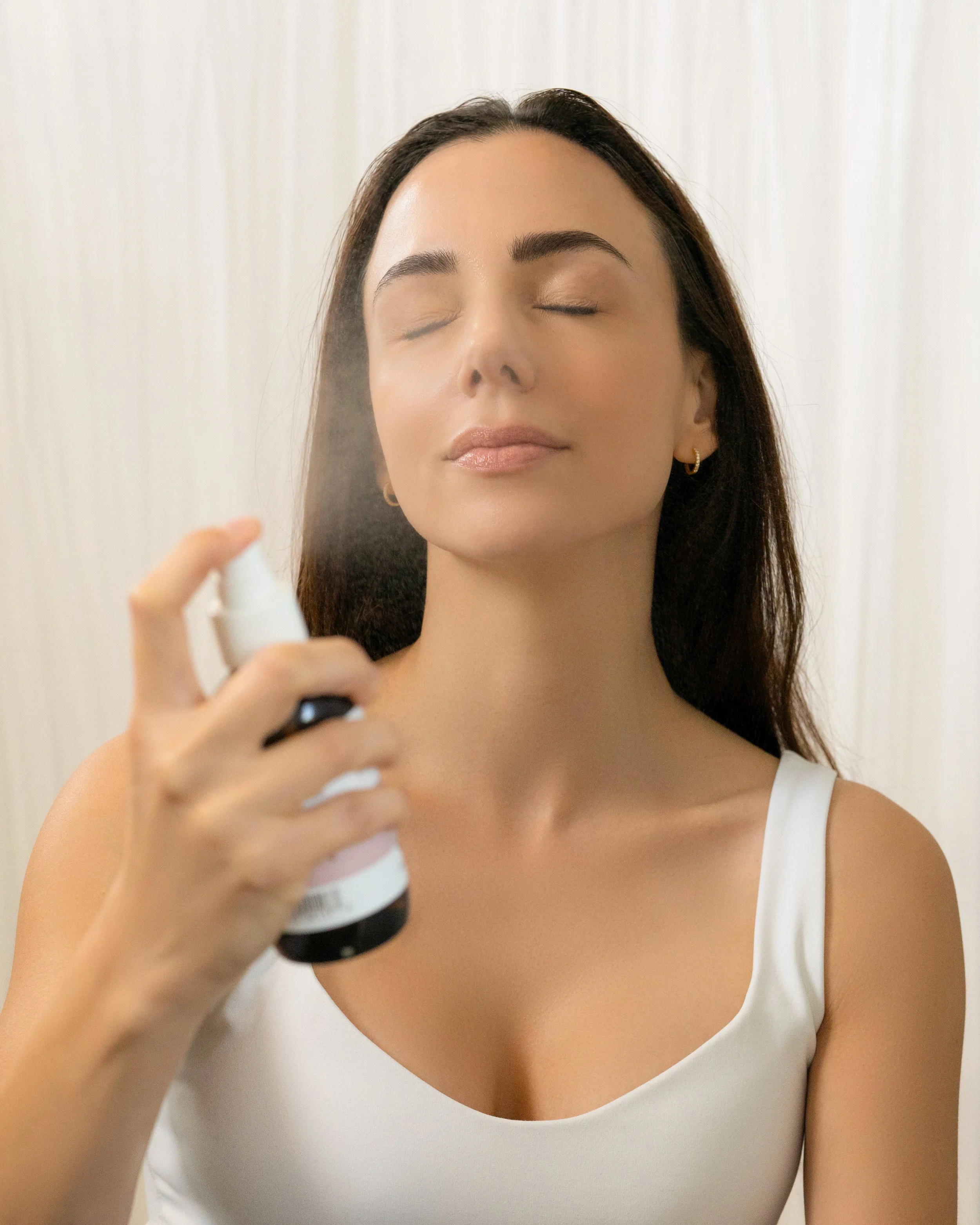 Studio skincare model with closed eyes spraying facial mist onto her face on a cream curtain backdrop.