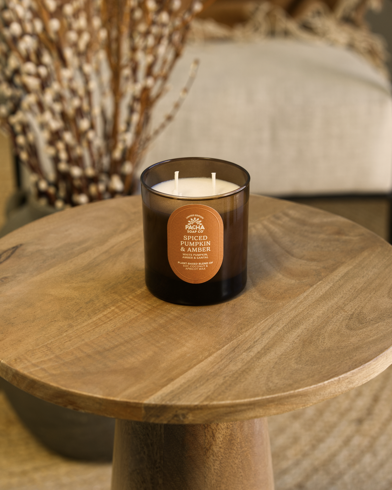Elevated lifestyle product photography of a Spiced Pumpkin & Amber scented candle in an amber glass jar on a wooden table with plant elements in a luxury home setting.