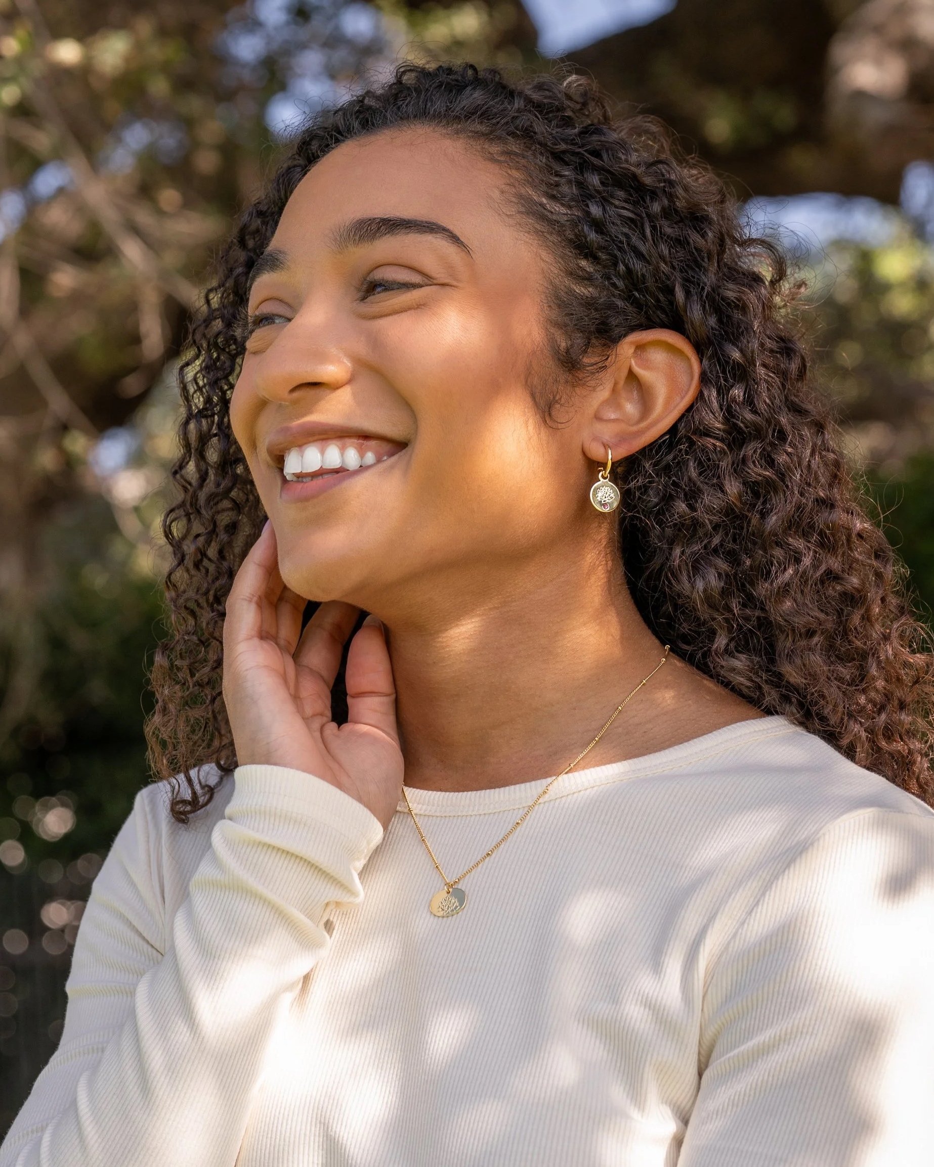 Elevated outdoor lifestyle photography of on-model jewelry with a smiling woman with curly hair wearing gold jewelry and a white top outdoors.