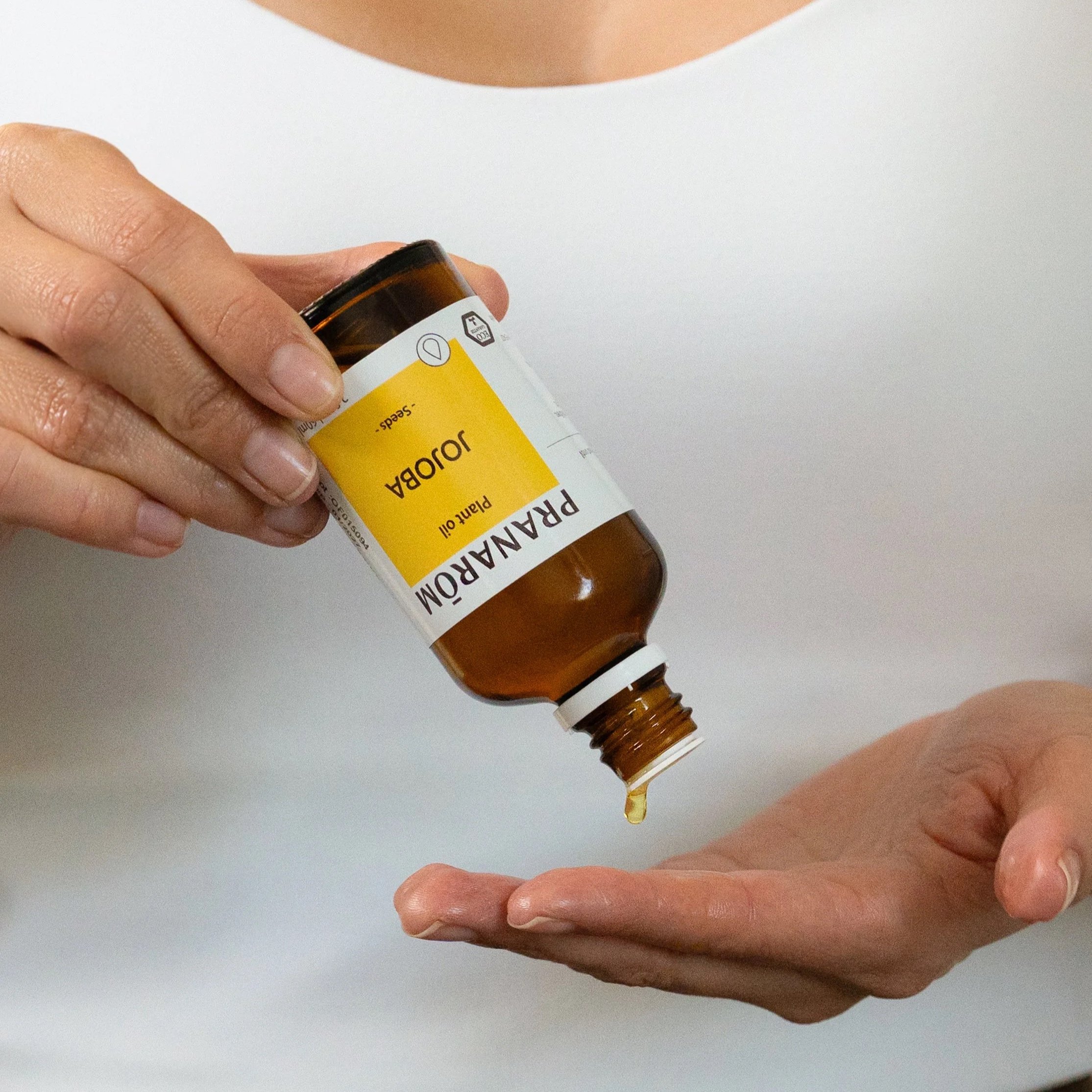Luxury skincare product photography of a hand model applying Jojoba plant oil from an amber bottle to their hand in a clean studio setting.