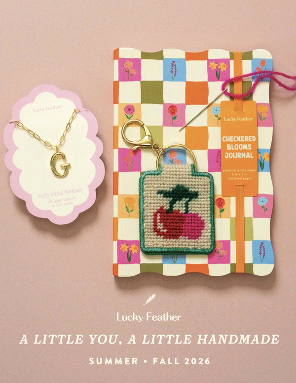 Catalog photography of handmade accessories featuring a gold initial G necklace and embroidered red cherry keychain on a colorful checkered floral journal.