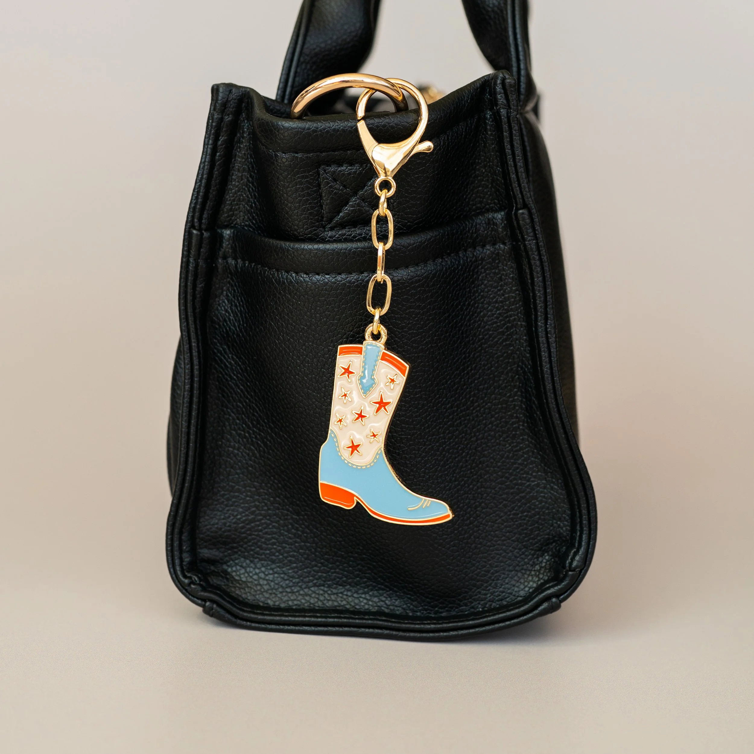Elevated studio product photography of a black leather handbag with a gold bag charm featuring a colorful cowboy boot bag charm.