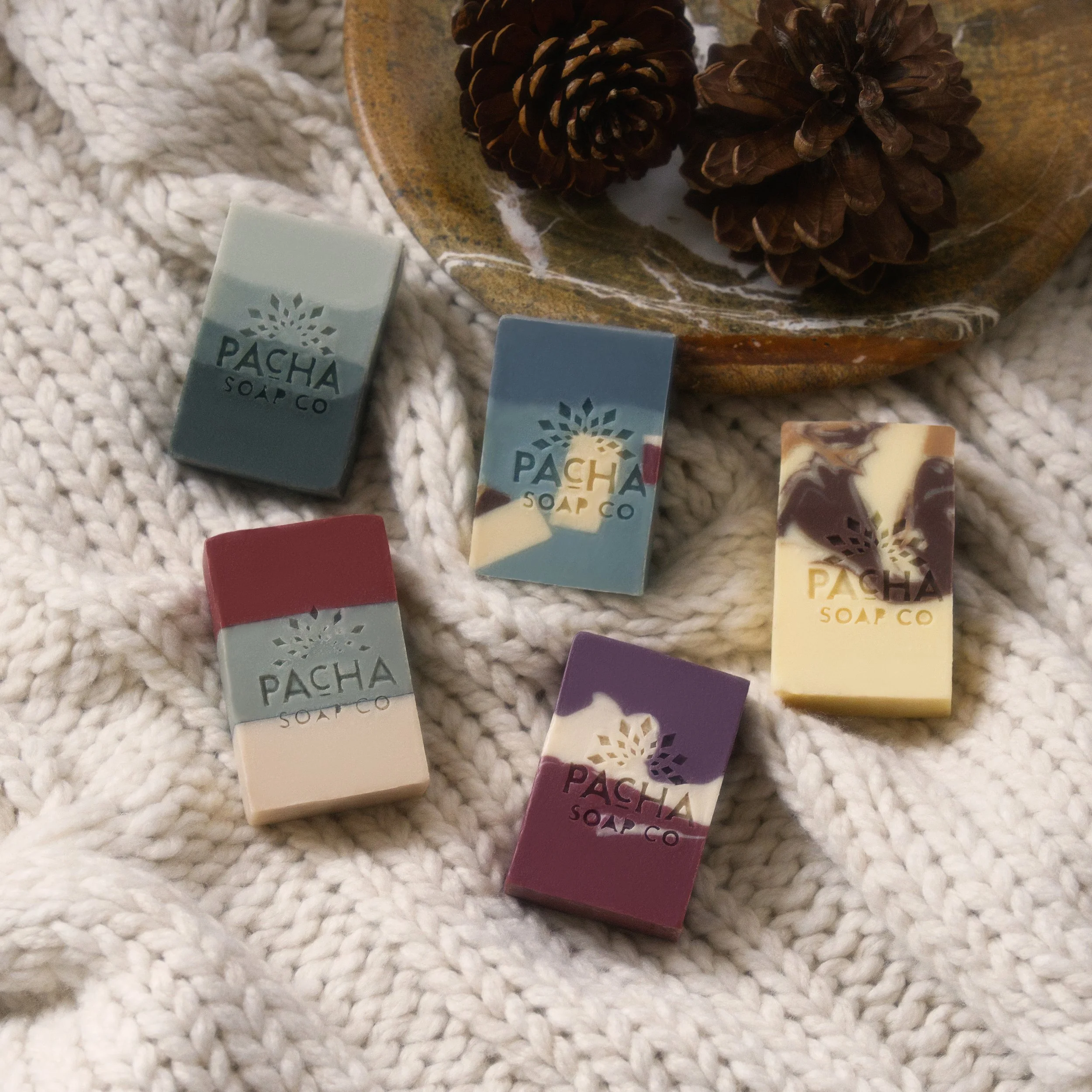 Elevated product photography of a colorful handmade soap bars arranged on a white knitted blanket beside a rustic dish of pinecones in a luxury holiday home setting.