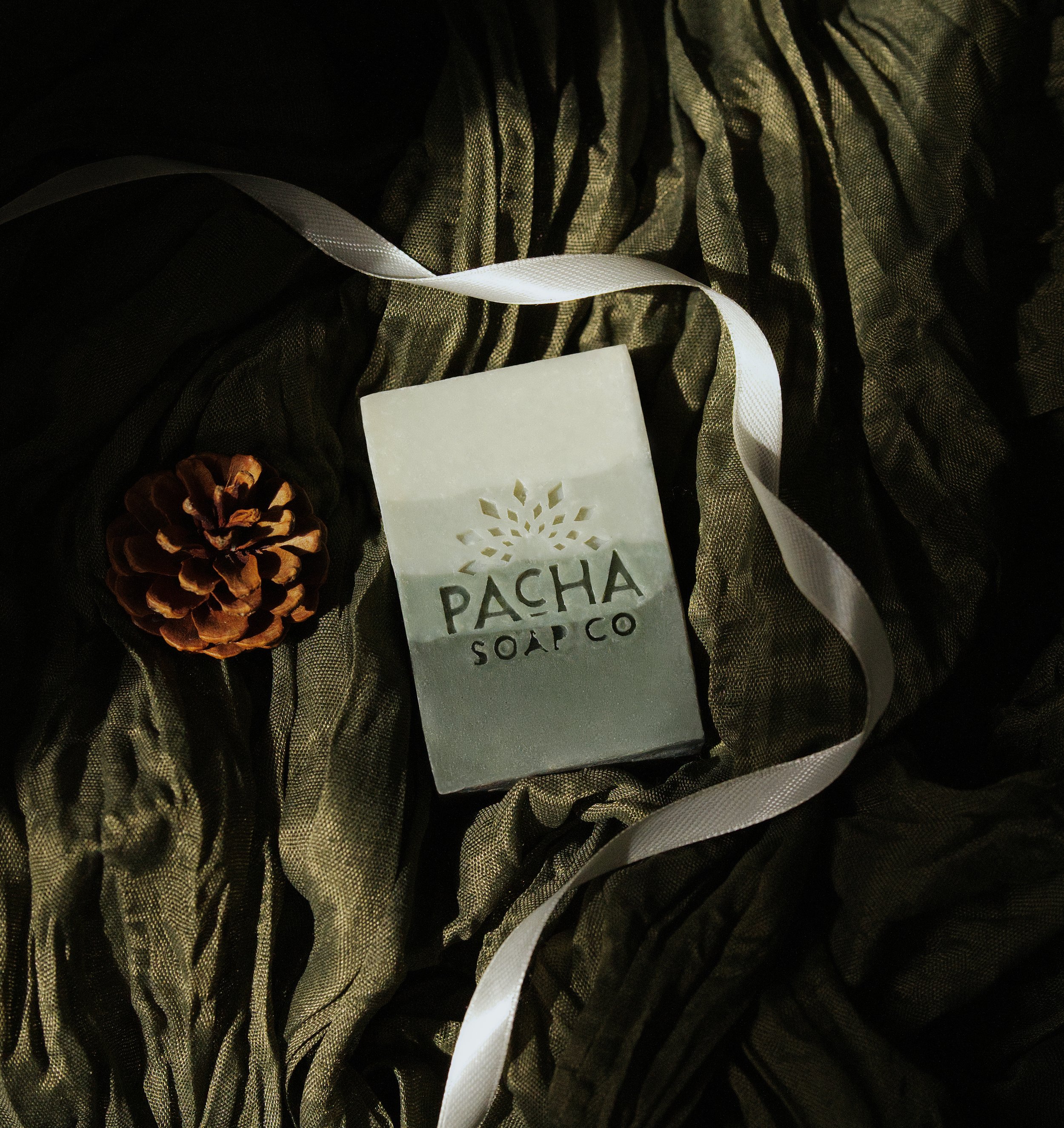 Elevated e-commerce photography of a handmade holiday soap bar with snowflake design, white ribbon, and pinecone on dark green fabric.
