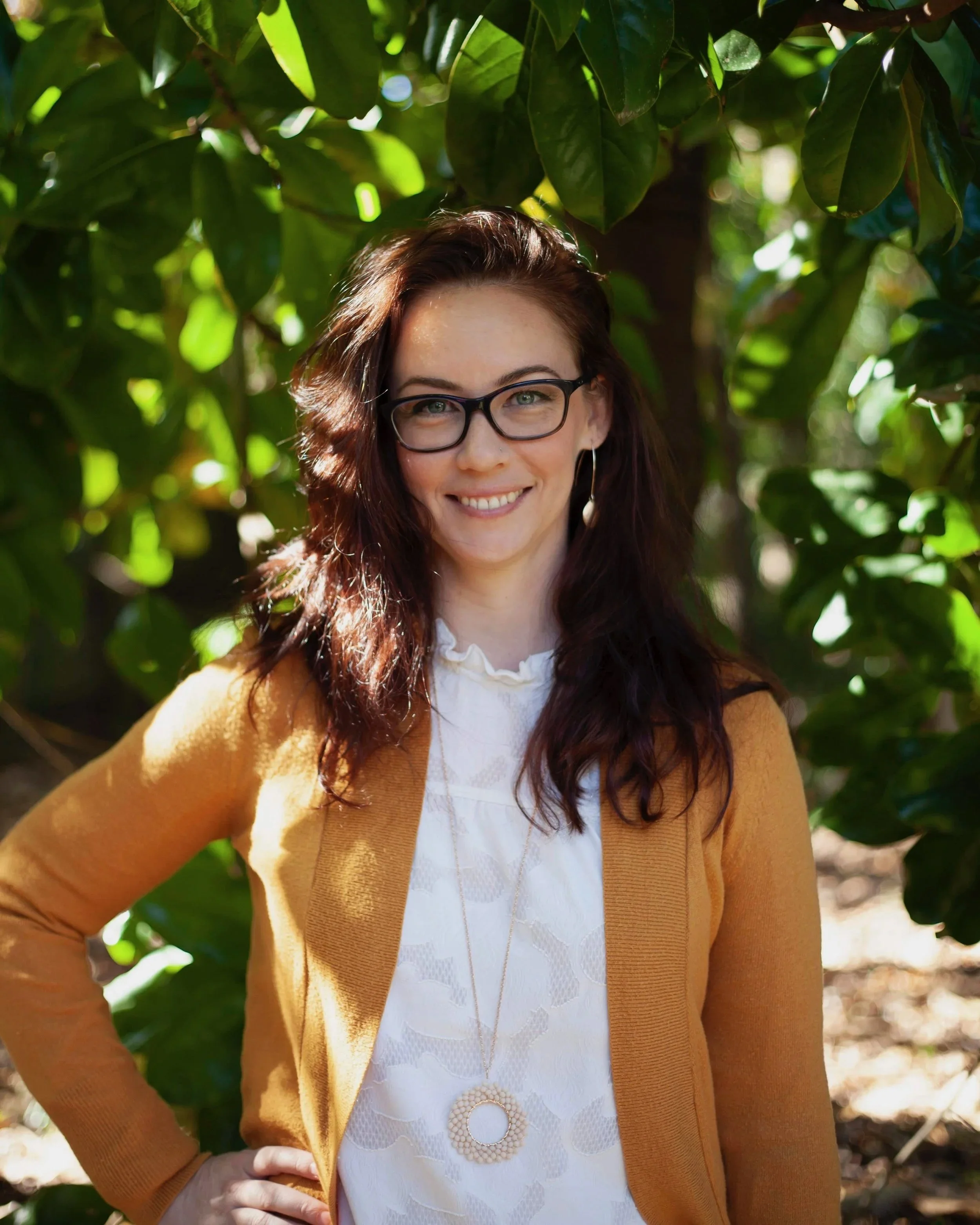 A woman with long brown hair, glasses, and hoop earrings, smiling, wearing a white blouse and a brown cardigan, standing outdoors among green leafy trees.