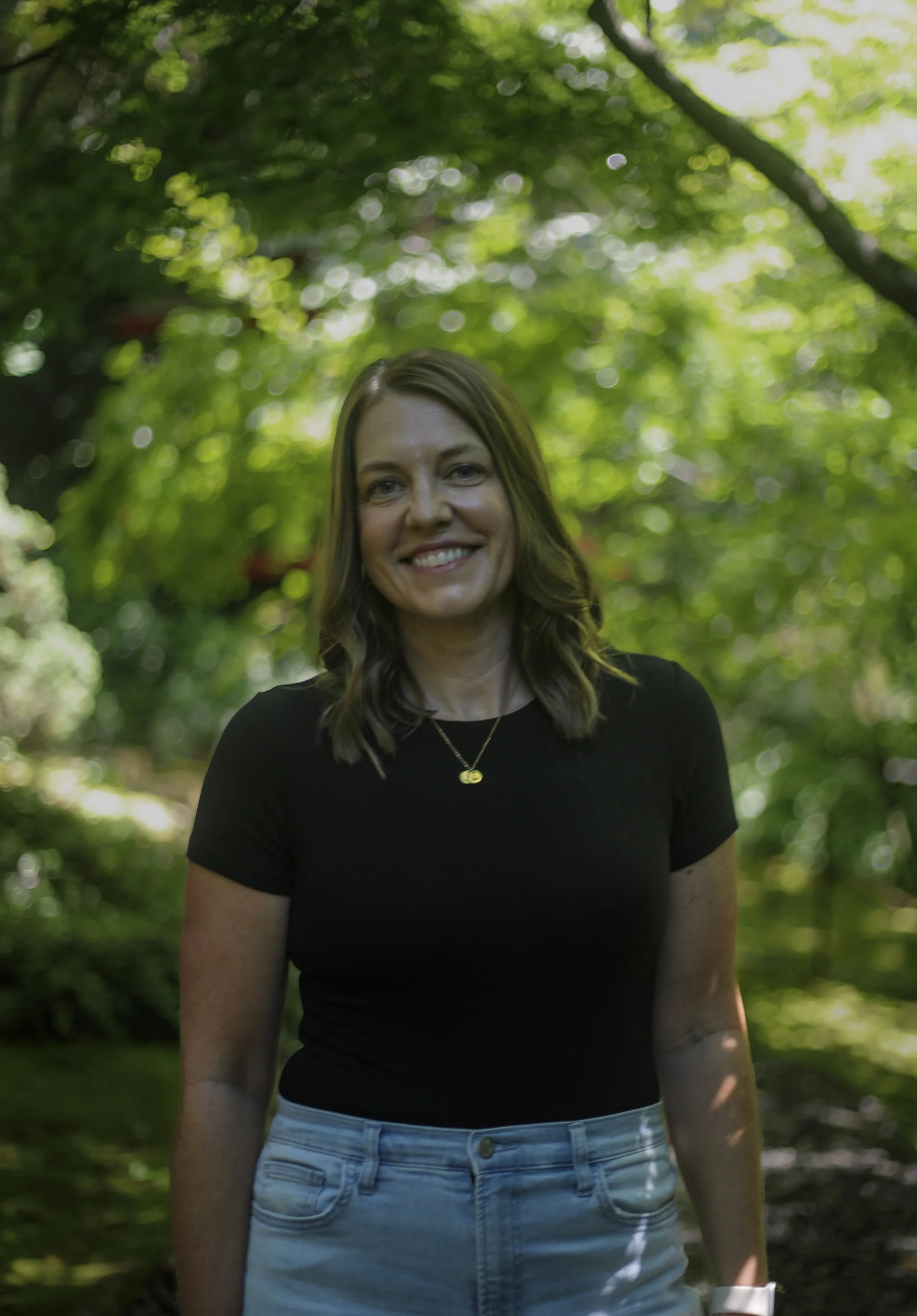 A woman with long brown hair smiling outdoors in a green, leafy environment wearing a black t-shirt and light-colored jeans.
