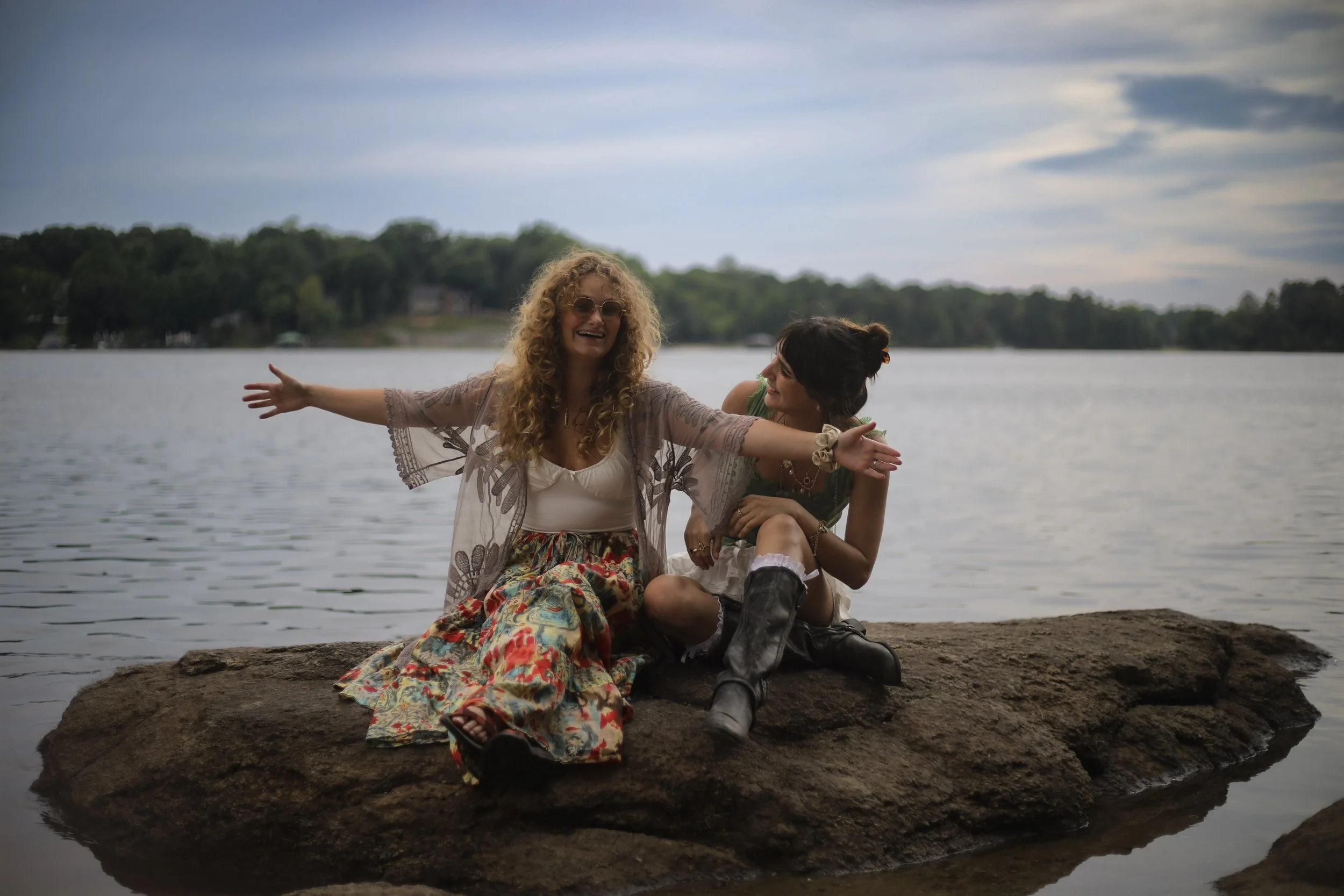 Two women sitting on a rock by the water, smiling and having fun. One woman with curly hair and sunglasses has her arms outstretched. The other woman has dark hair in a bun and is wearing boots, looking at her friend. There are trees and a cloudy sky