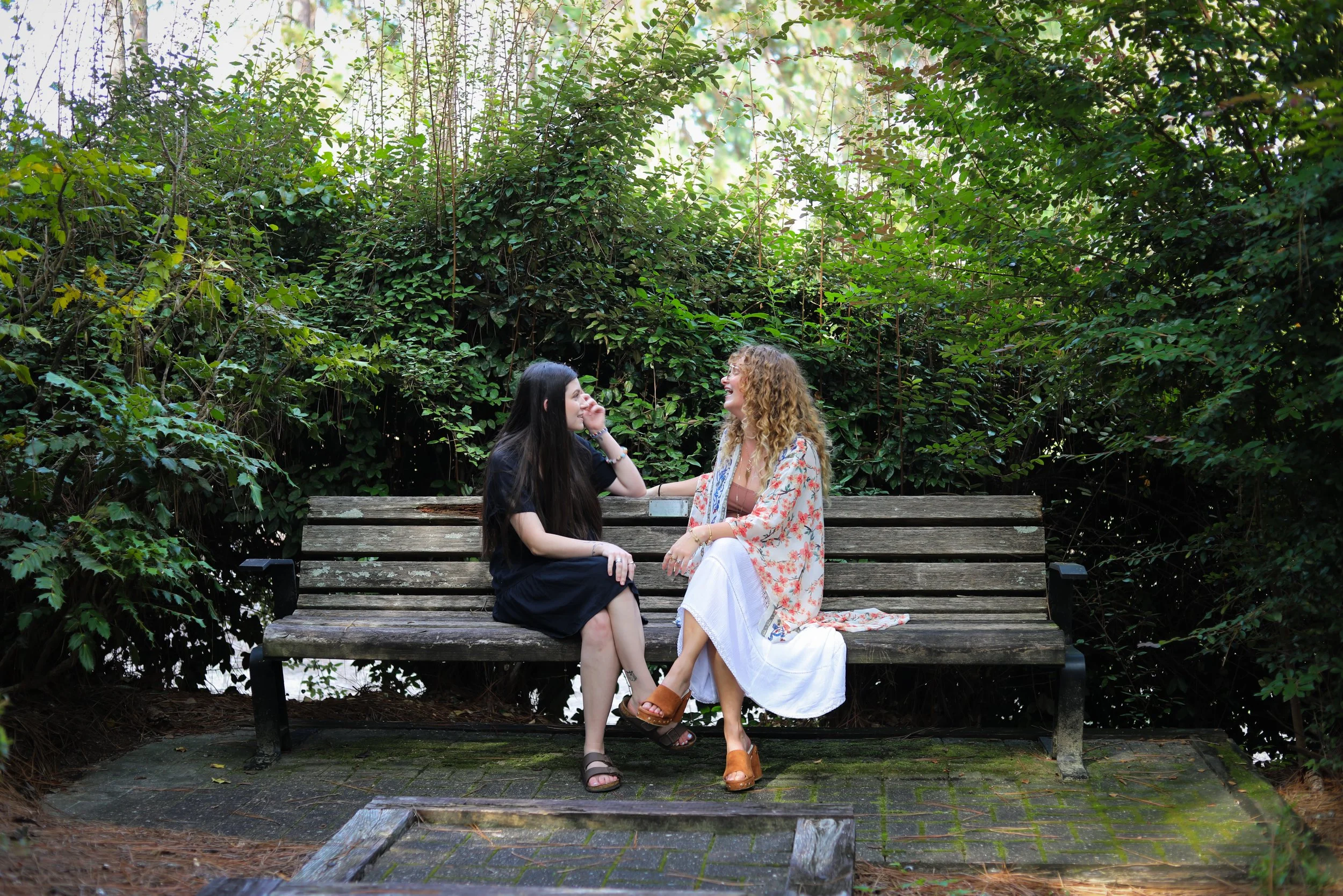 Two women sitting on a wooden park bench surrounded by green bushes, engaged in conversation and smiling.