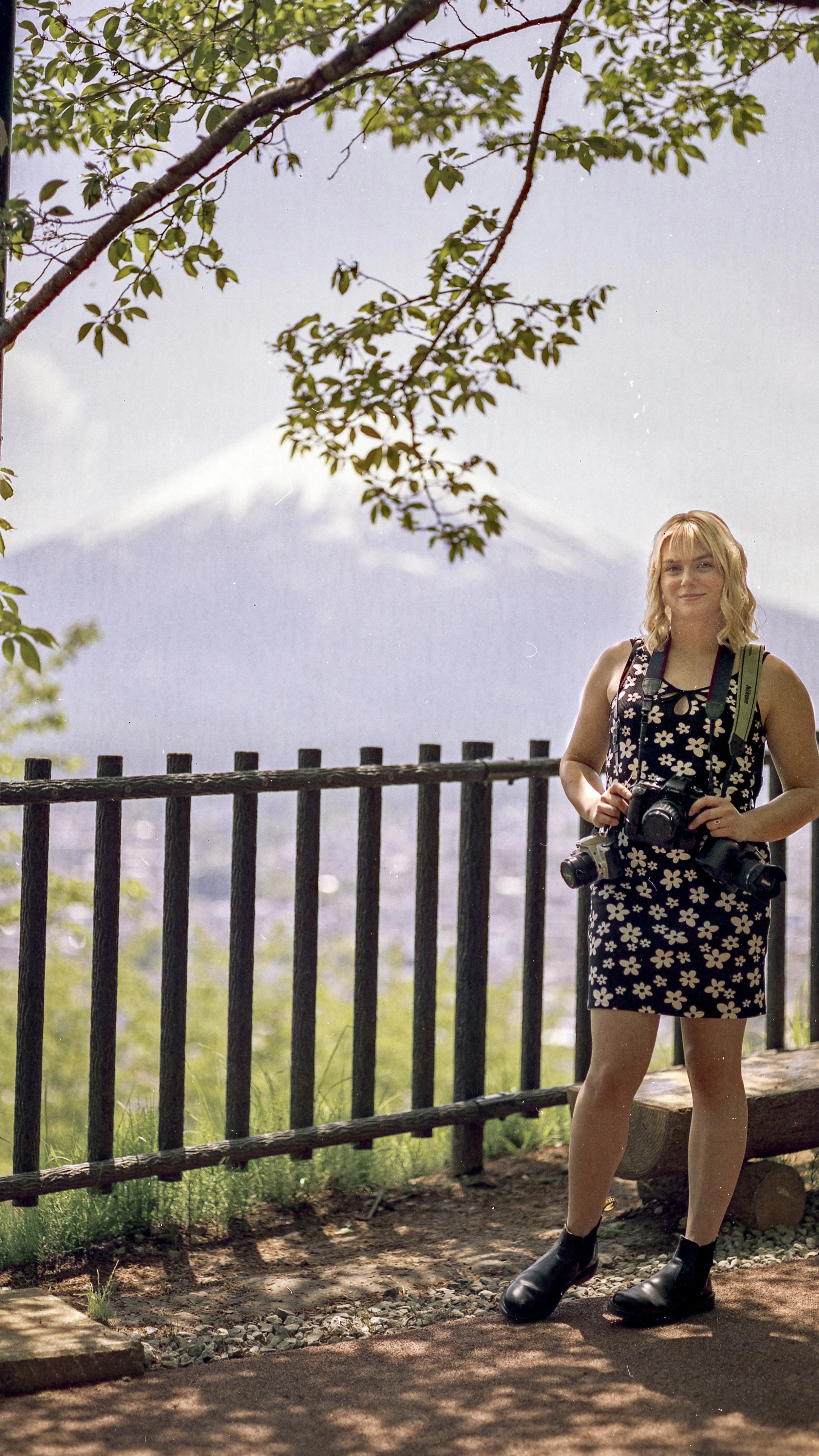 A woman with blonde hair wearing a flowered dress and black boots standing outdoors, holding two cameras with a scenic mountain view in the background.