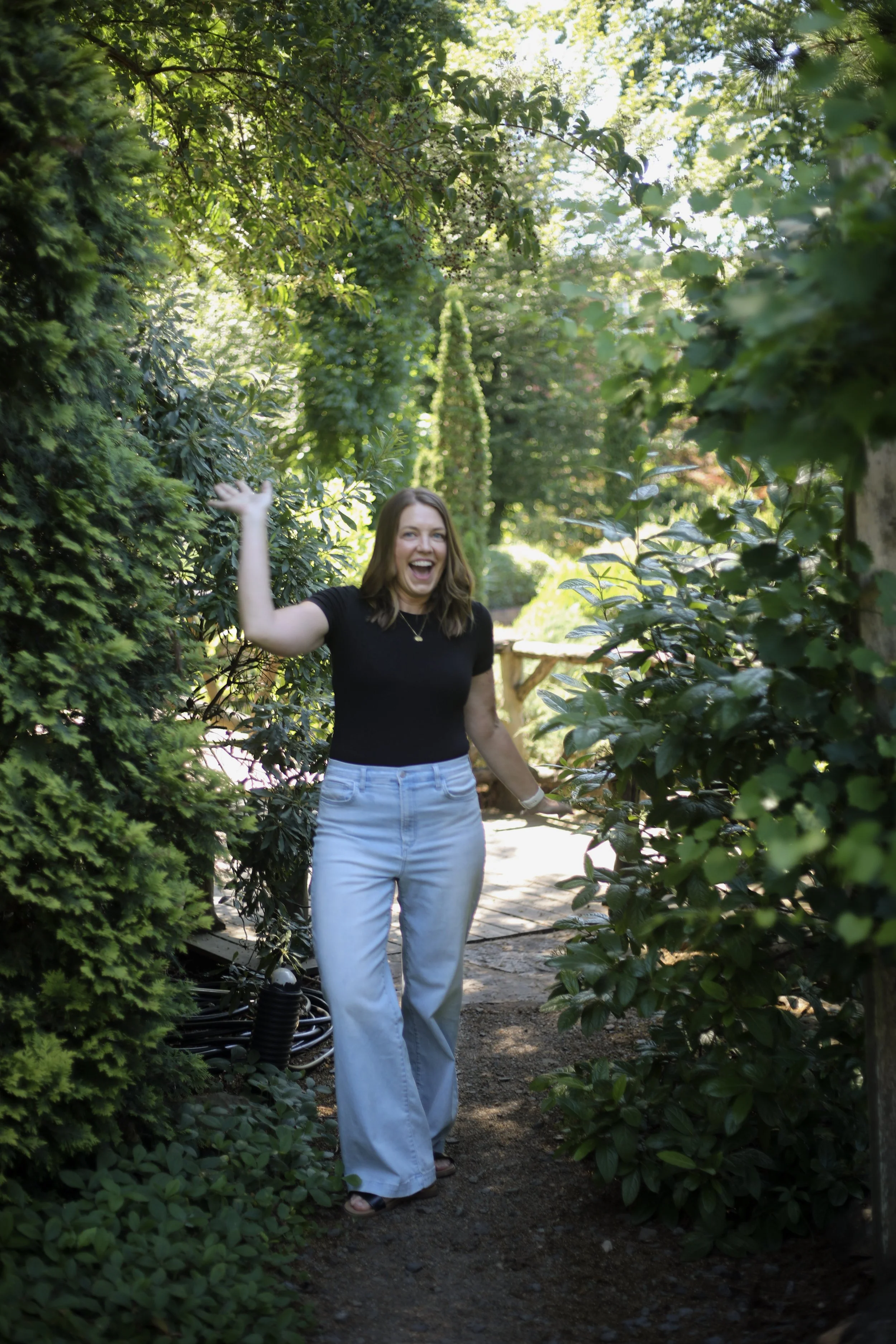 A woman in a black top and light blue jeans smiling and waving while standing in a lush garden pathway surrounded by green foliage.
