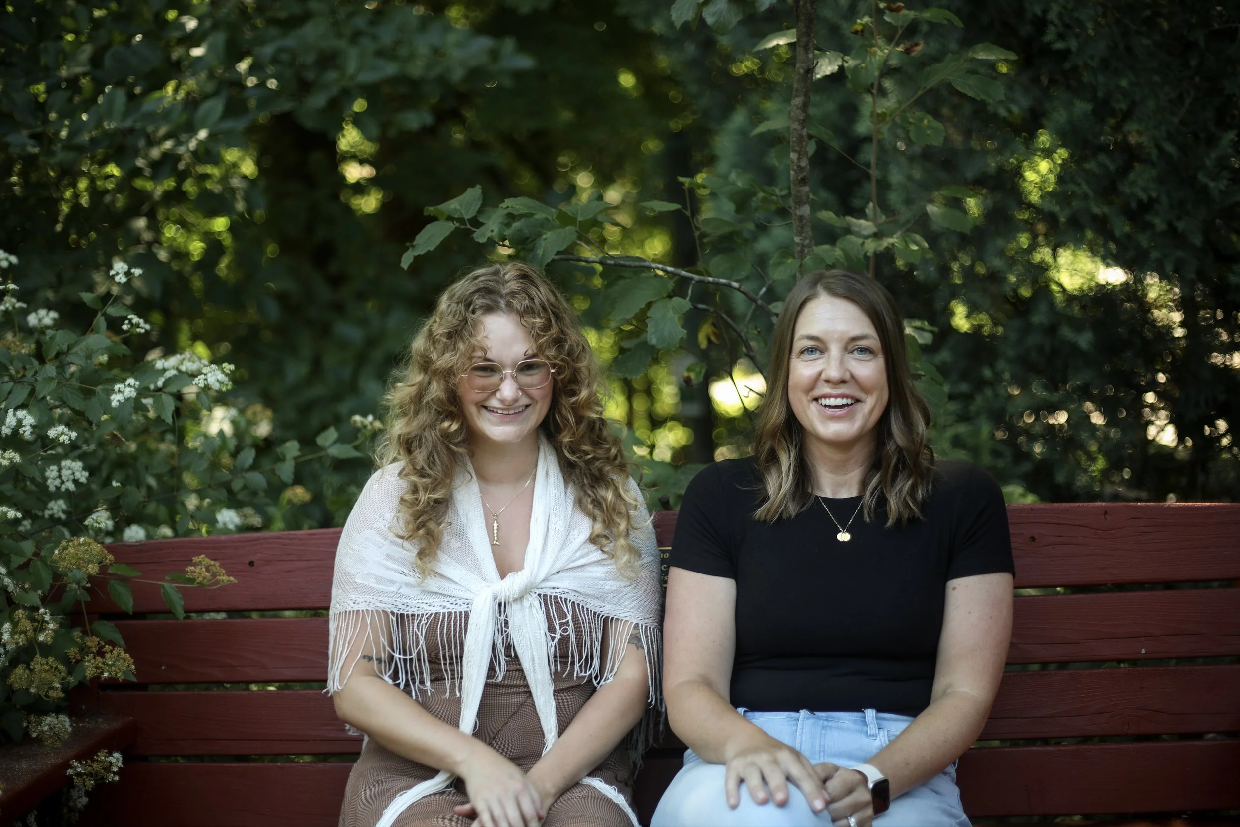 Two women sitting on a red wooden bench in a lush, green outdoor setting, smiling at the camera.
