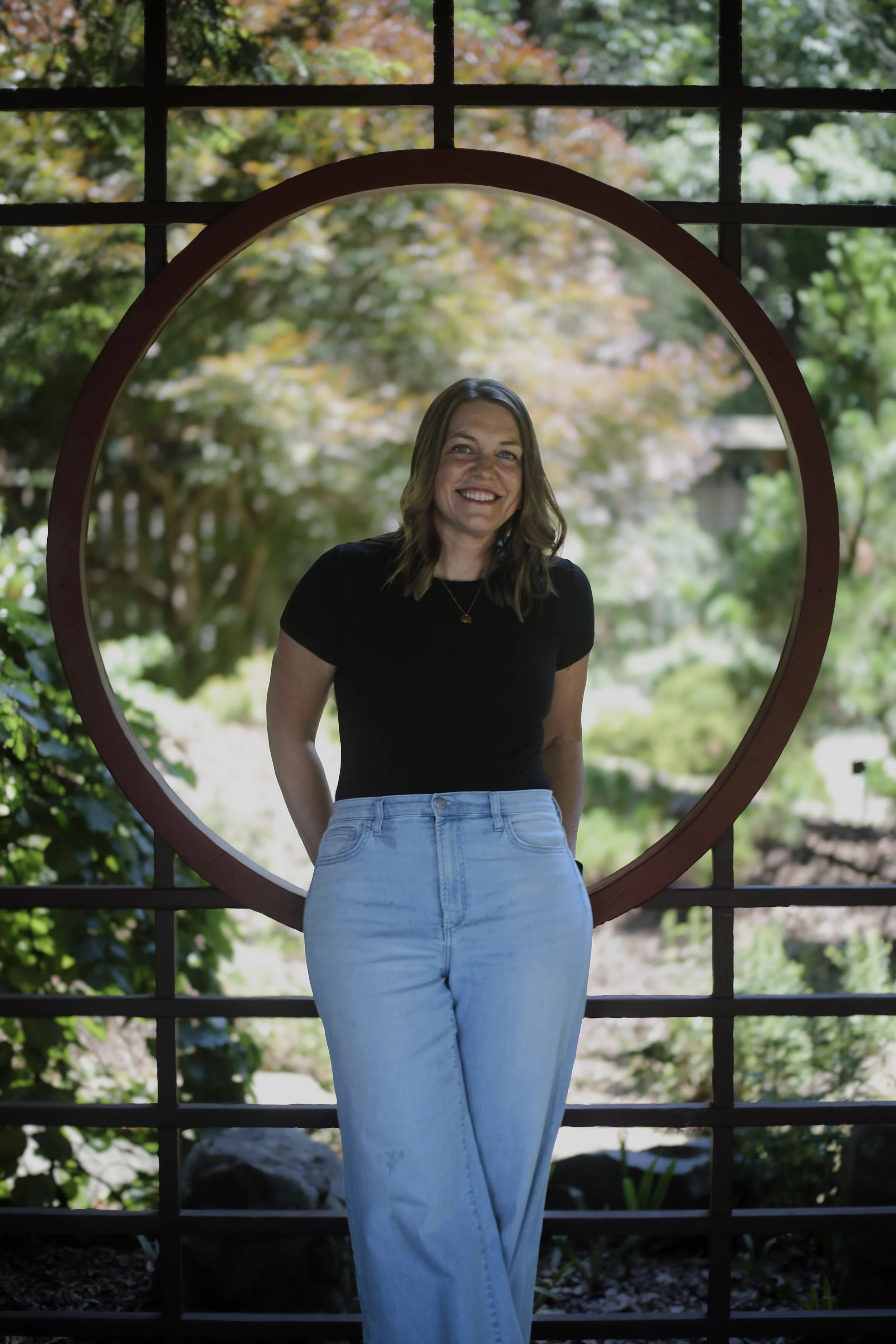 A woman with shoulder-length brown hair and a black top smiling, standing in front of a decorative round mirror with a garden and trees reflected behind her.