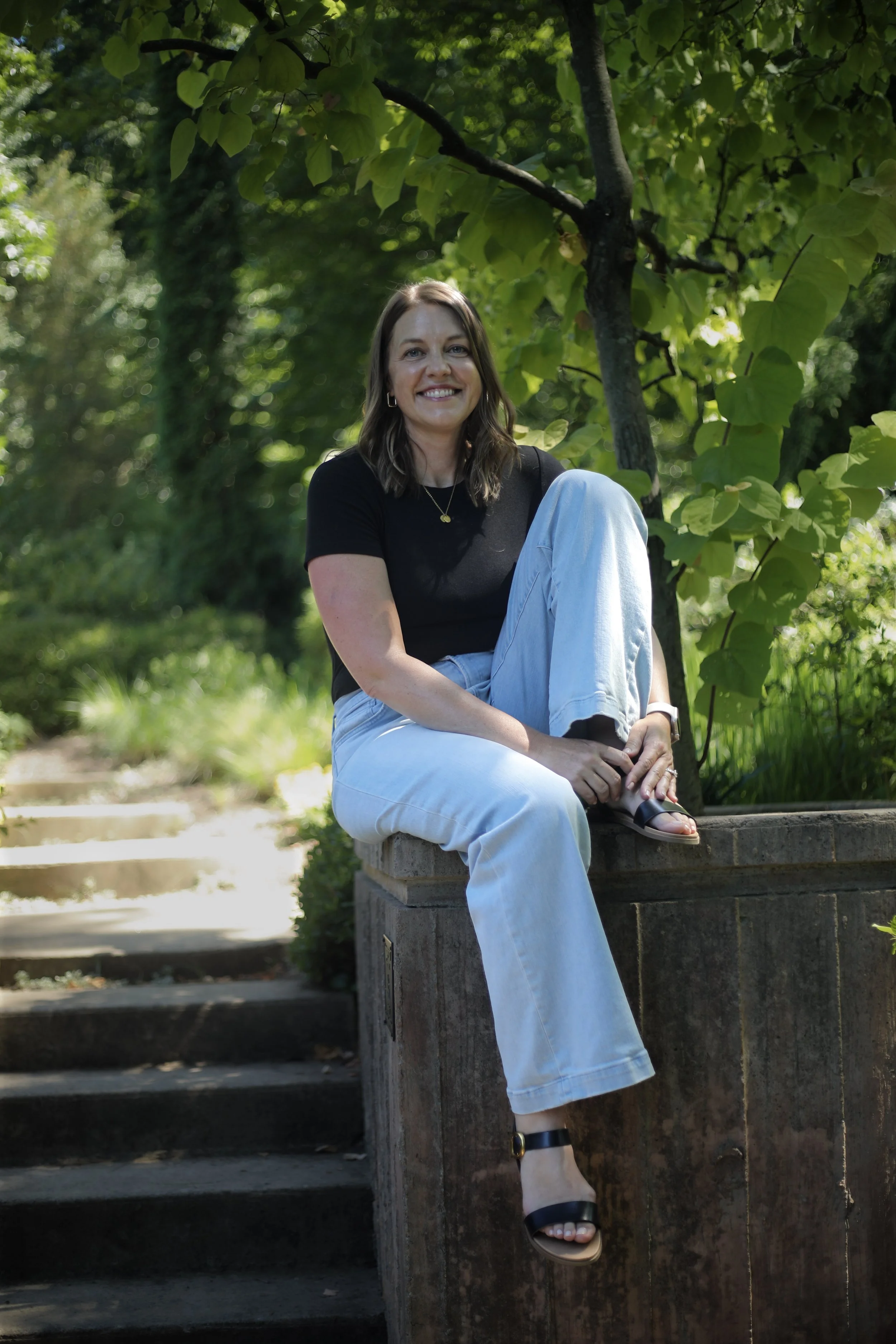 A woman sitting on a stone ledge outdoors surrounded by green trees and foliage, smiling at the camera with relaxed posture, wearing a black t-shirt, white jeans, and black sandals.