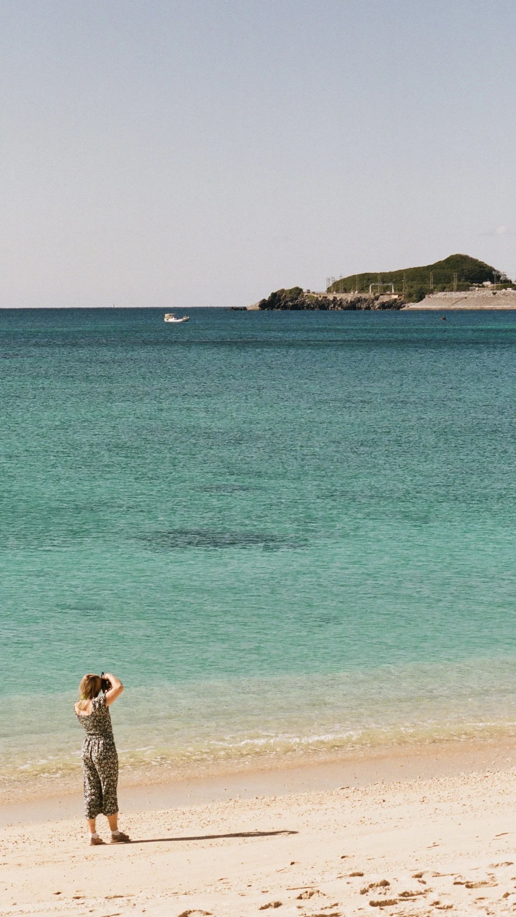 A woman standing on a sandy beach taking a photograph of the ocean with hills in the distance.