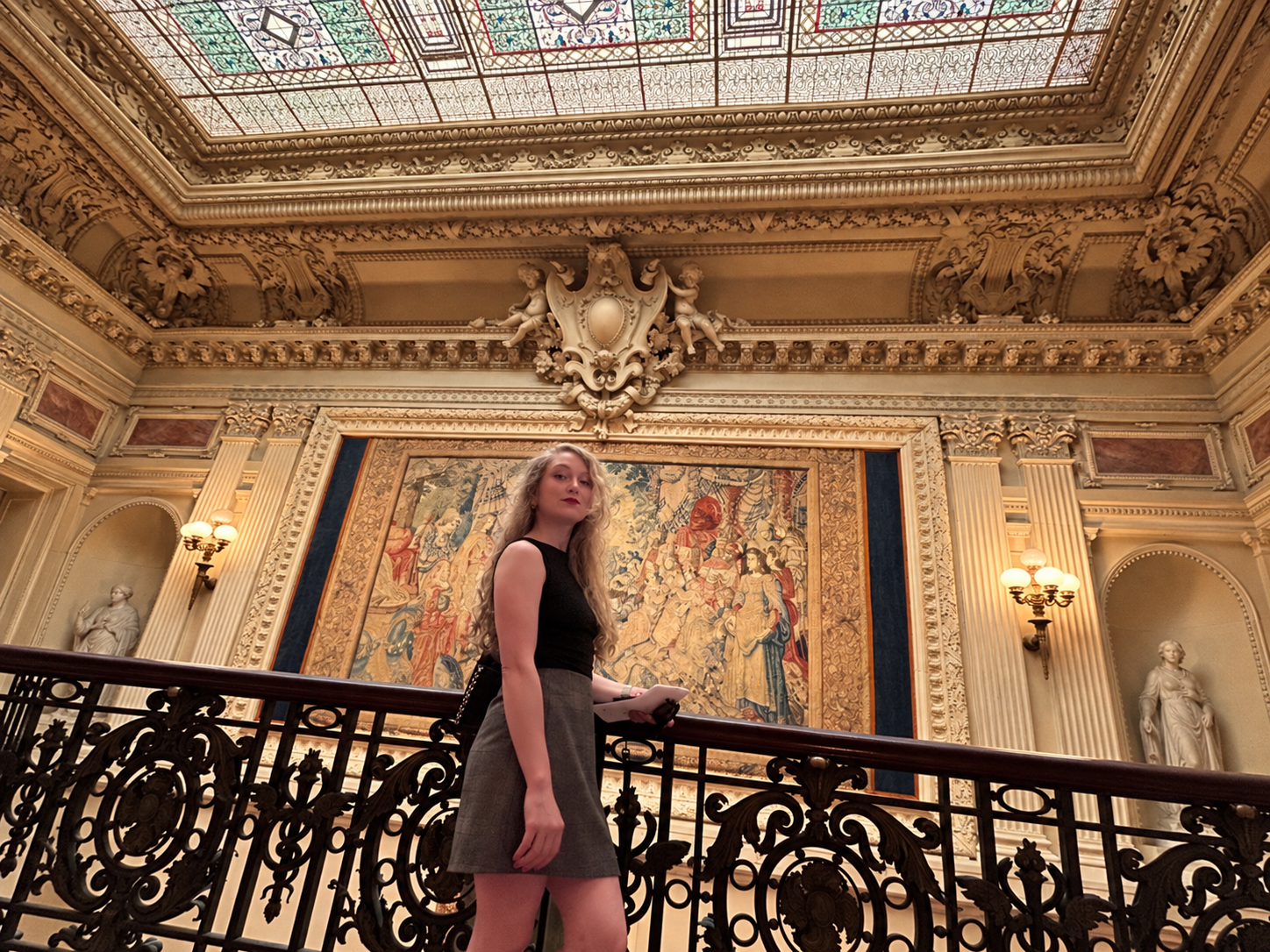 Young woman with long blonde hair standing on a staircase inside an ornate, classical-style room with elaborate molding, statues, and a large tapestry on the wall behind her.