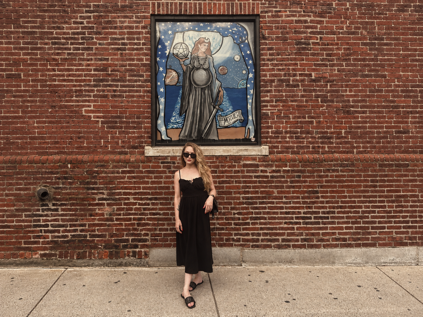 A woman in a black dress and sunglasses stands in front of a red brick wall with a mural of the Lady of the Lake from Arthurian legend holding a sword and a chalice, with celestial background and a banner that says 'the Mother'.
