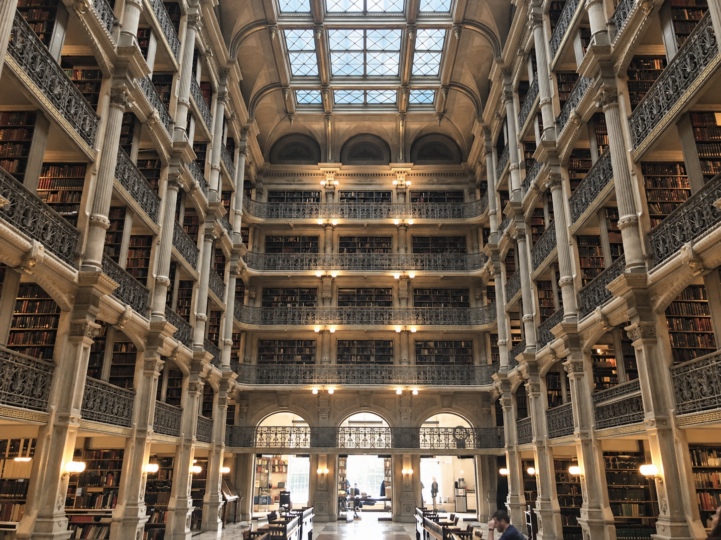Interior of a grand multi-level historic library with ornate iron balconies, large arched windows, and a glass ceiling letting in natural light.