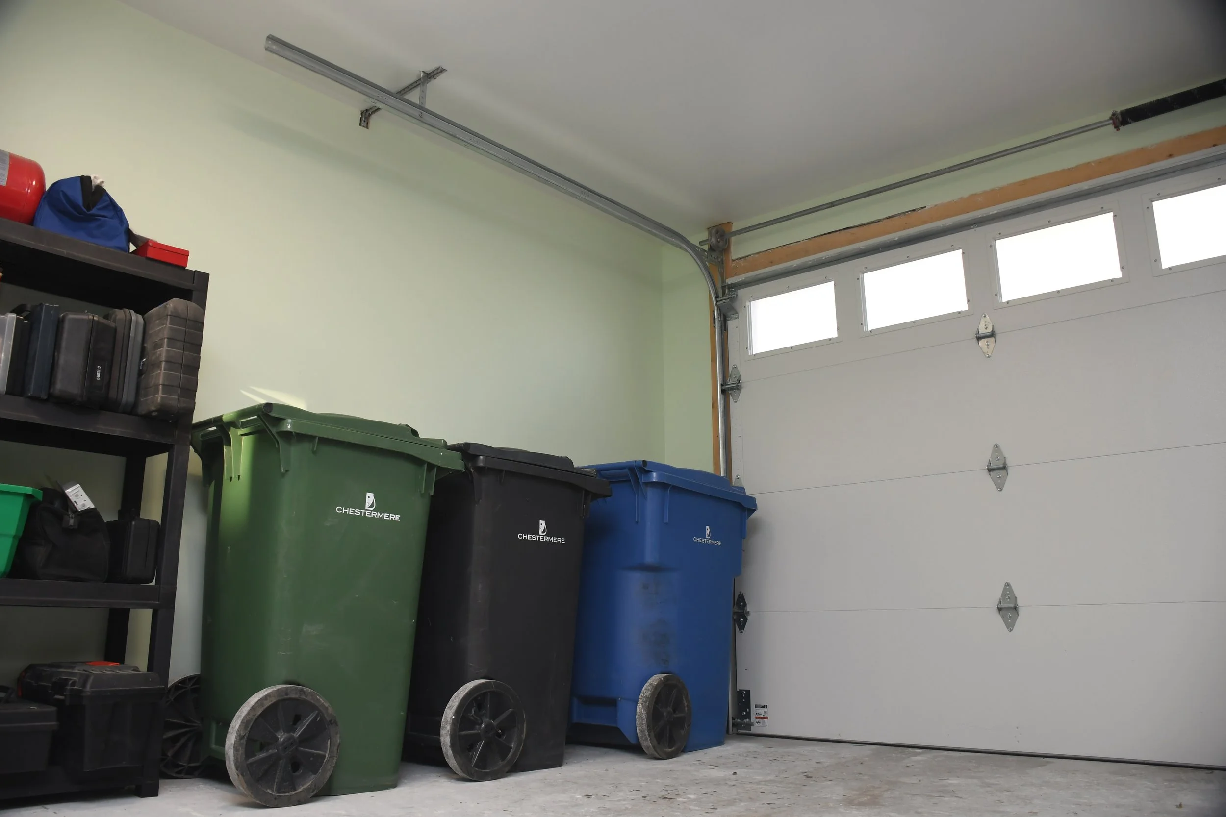 Garage with three trash bins in green, black, and blue, and a black shelving unit with tools and containers, garage door with windows.