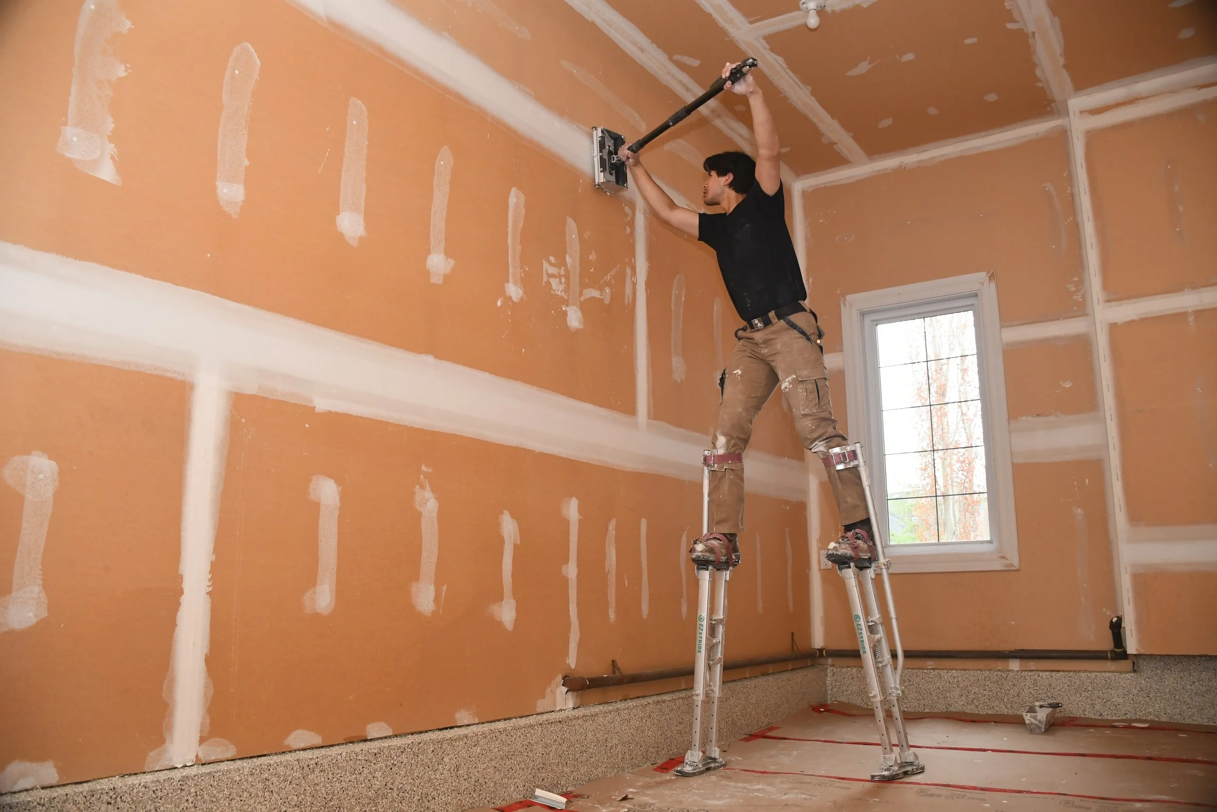 A man standing on a ladder in a room with unfinished drywall, holding a sander and working on the ceiling.