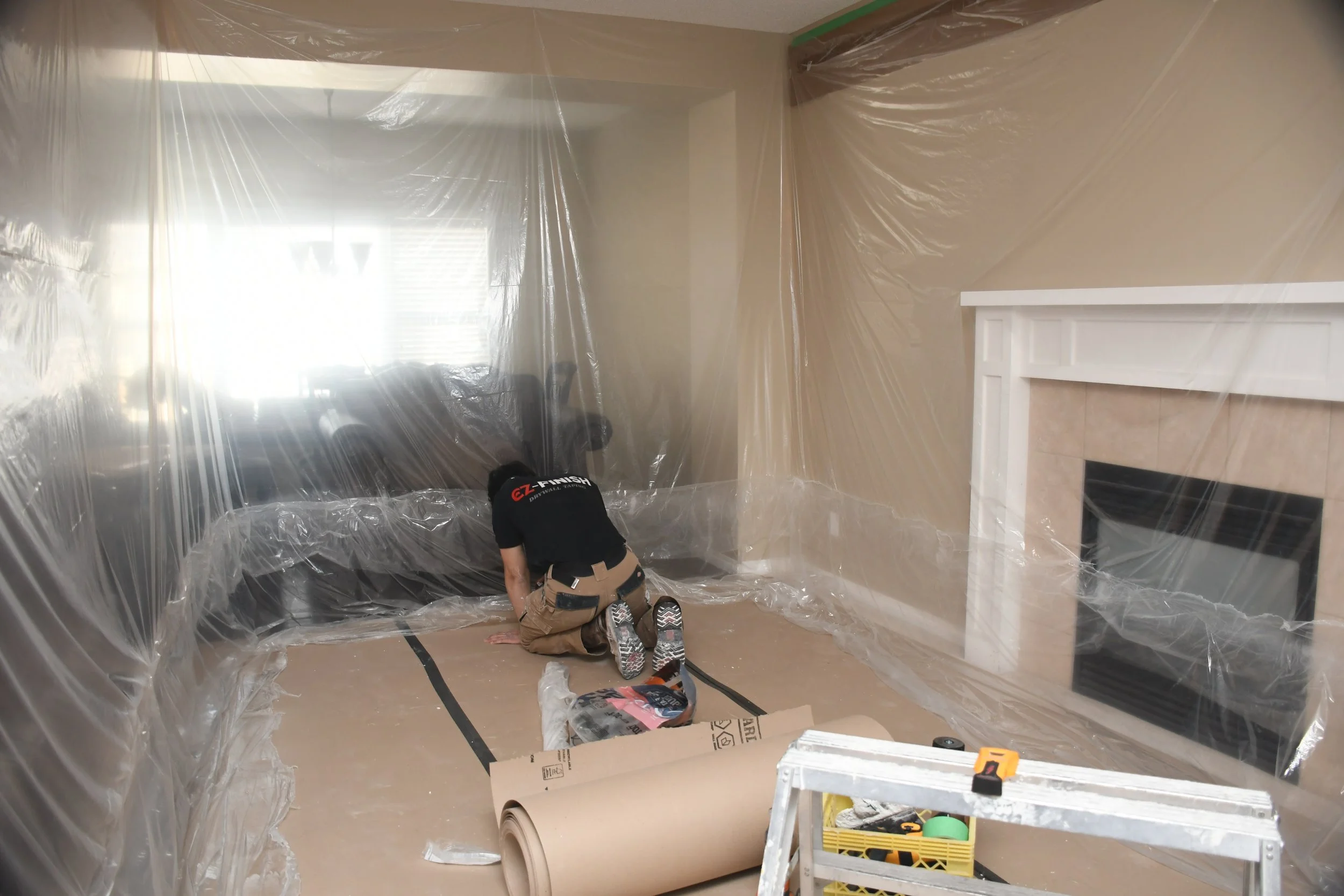 A person working on a room renovation, kneeling on the floor, surrounded by plastic sheeting, wallpaper, and tools near a fireplace.