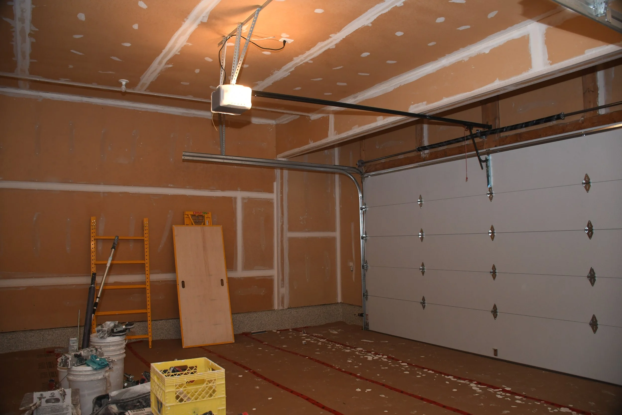 A garage interior under construction with unfinished drywall on the walls and ceiling, a closed sectional garage door on the right, and construction tools and supplies on the floor.