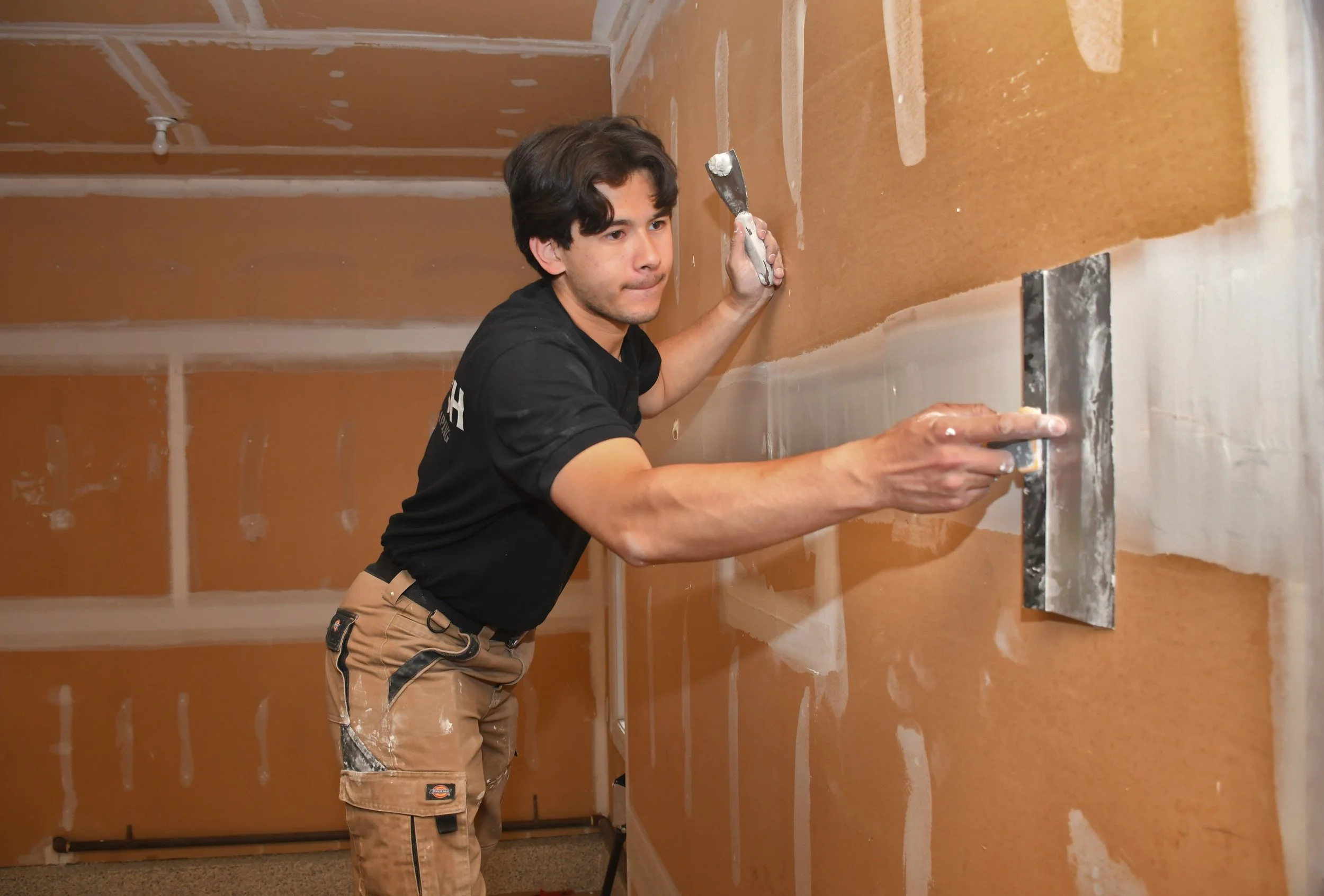 Man installing drywall in a room, using a level and trowel, with drywall seams showing.